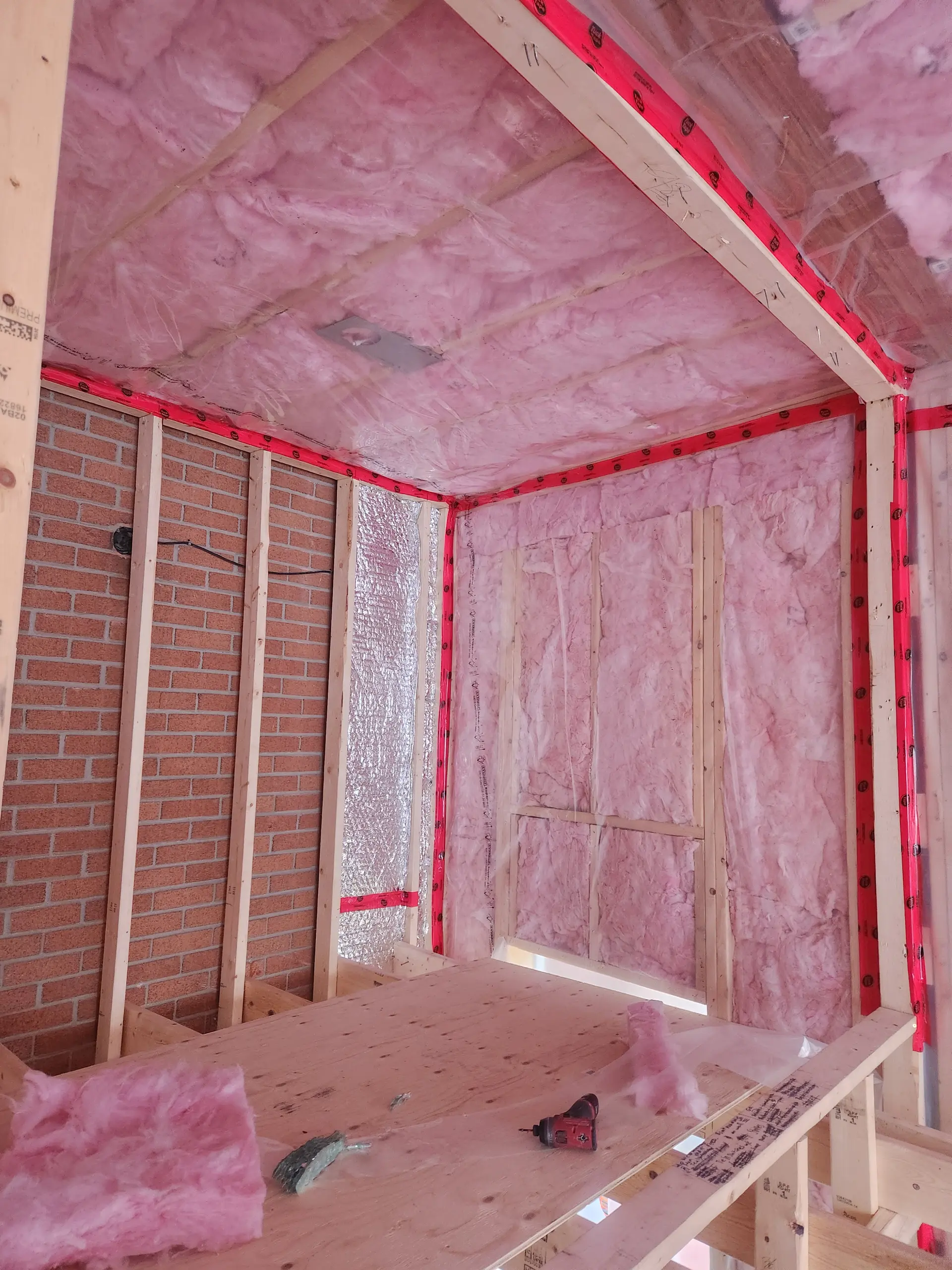 Pièce en construction avec isolant rose en fibre de verre, ossature de bois, pare-vapeur rouge et mur de brique intérieur