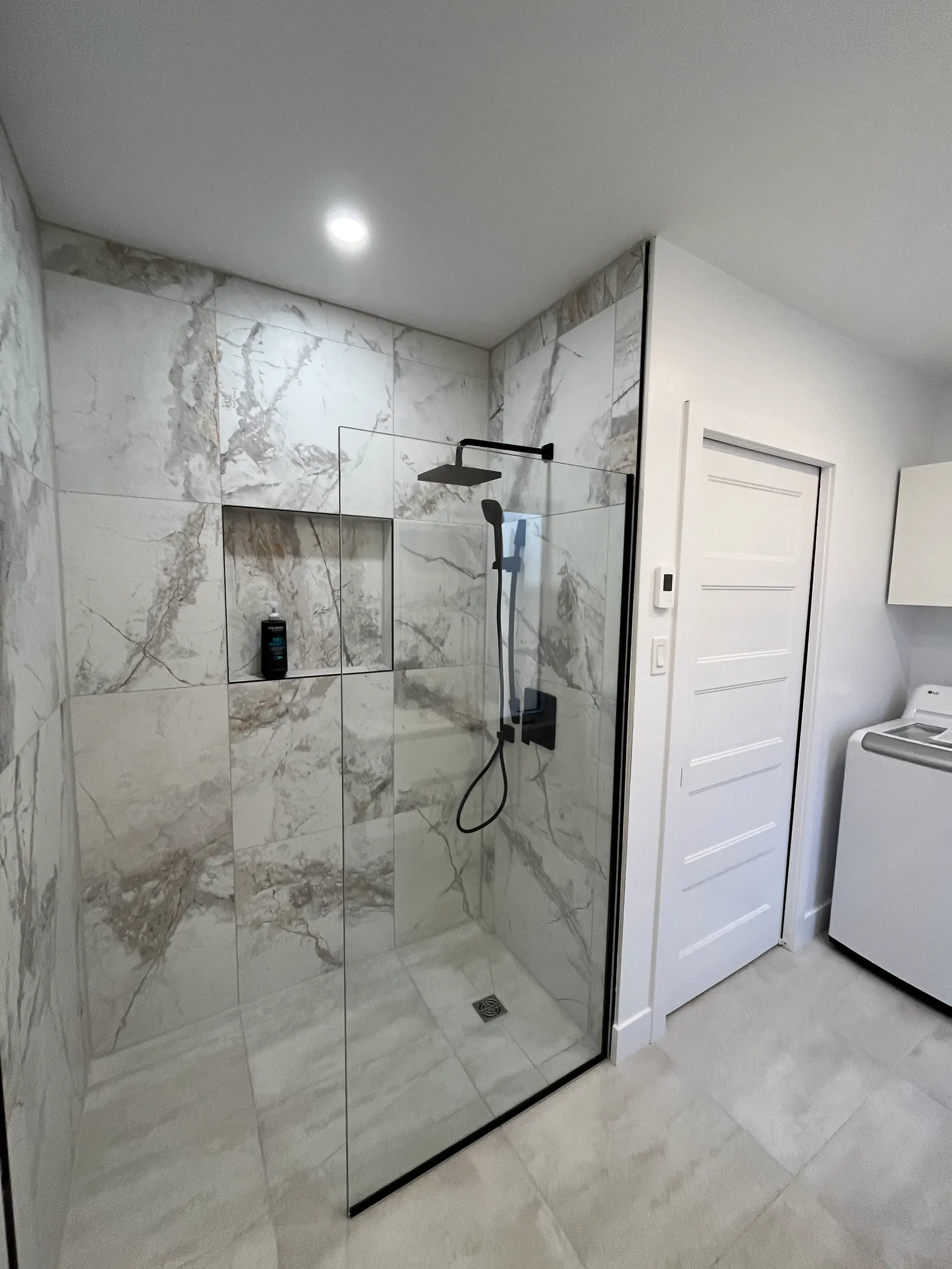 Douche italienne moderne avec grandes tuiles effet marbre, paroi de verre, robinetterie noire et niche murale intégrée dans une salle de bain lumineuse.