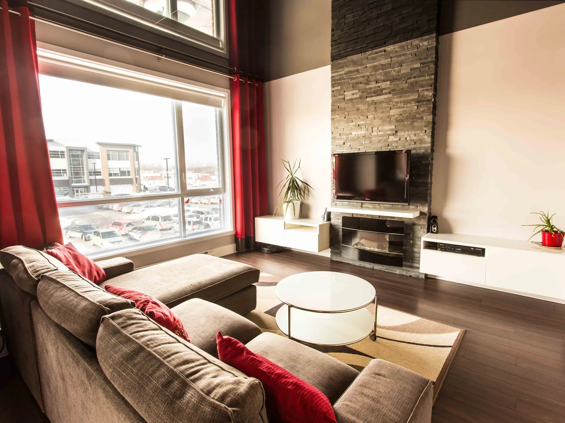 Bright modern living room with large window, red curtains, stone wall with fireplace and wall-mounted TV