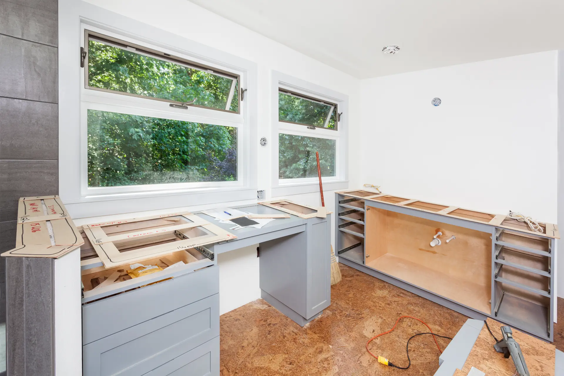 Cuisine en rénovation avec installation de nouveaux comptoirs et armoires grises sous des fenêtres lumineuses.