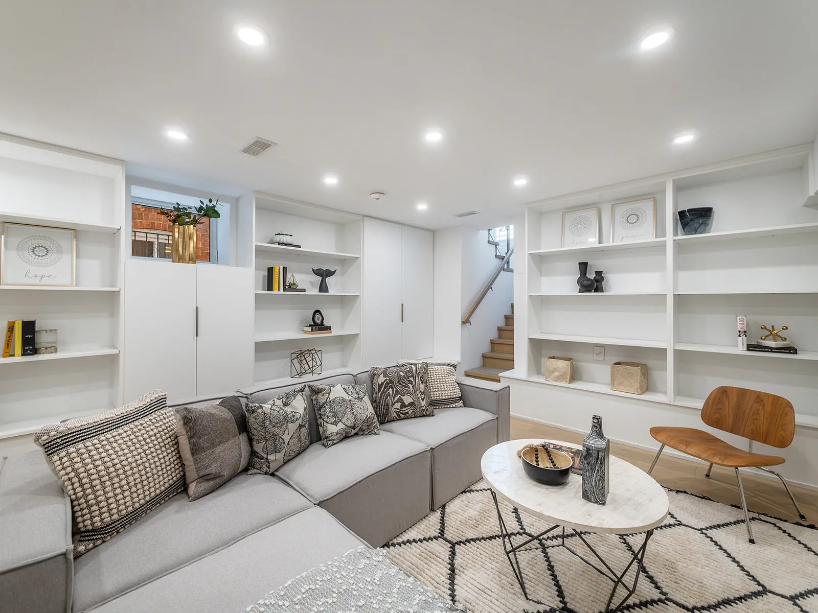 Sous-sol aménagé avec salon moderne comprenant un canapé modulaire, une bibliothèque intégrée, une table basse en marbre, un fauteuil en bois et une décoration minimaliste.