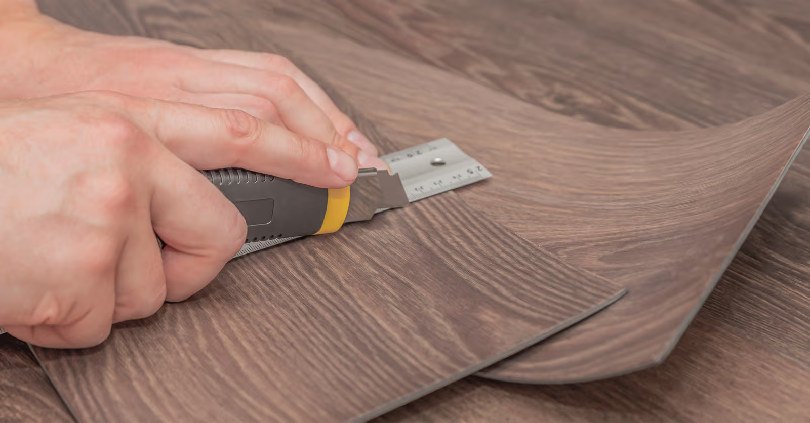 Main utilisant un couteau utilitaire et une règle pour découper une lame de plancher vinyle imitation bois lors d’une installation intérieure
