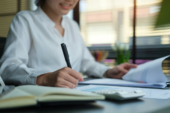 Professional woman taking notes and reviewing documents with a calculator on a modern office desk.