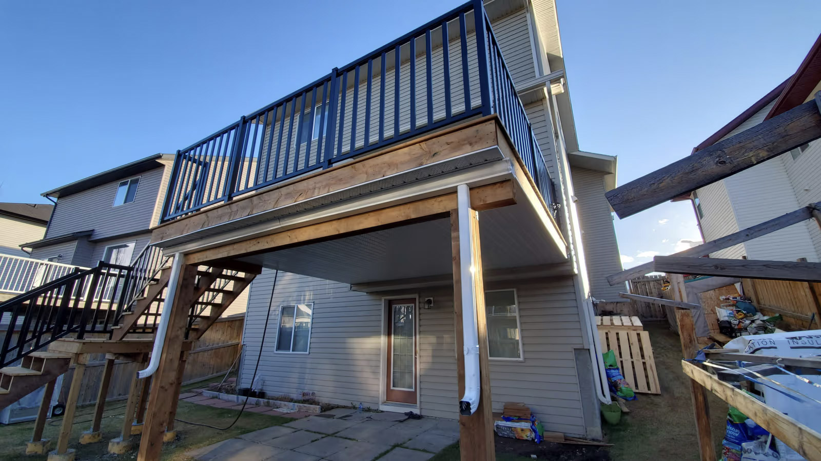 Balcon surélevé en bois avec garde-corps noir, escalier extérieur et structure sur poteaux derrière une maison résidentielle.
