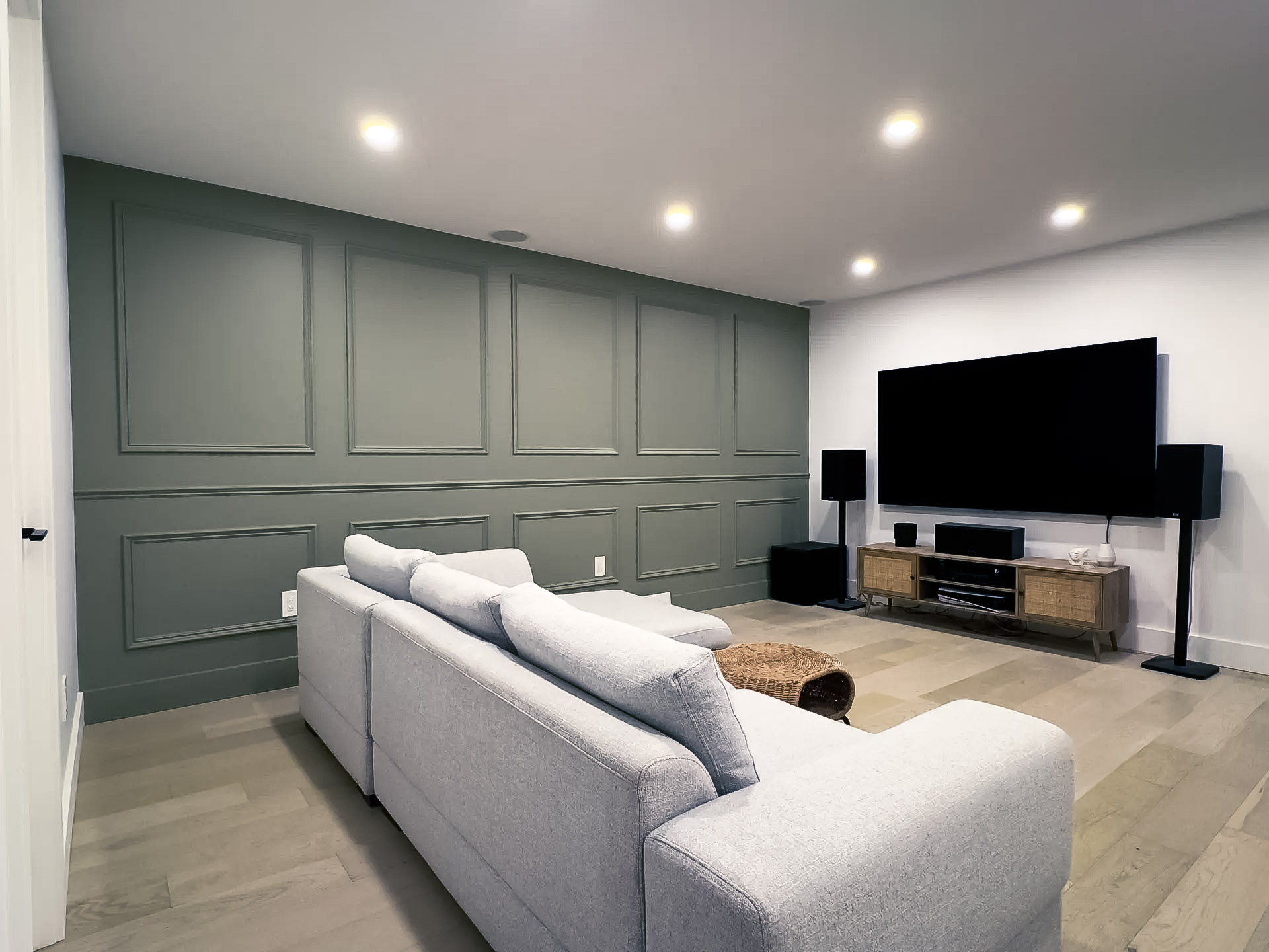 Modern home theater room with a light gray sofa, olive green paneled accent wall, large flat-screen TV, and surround sound system.