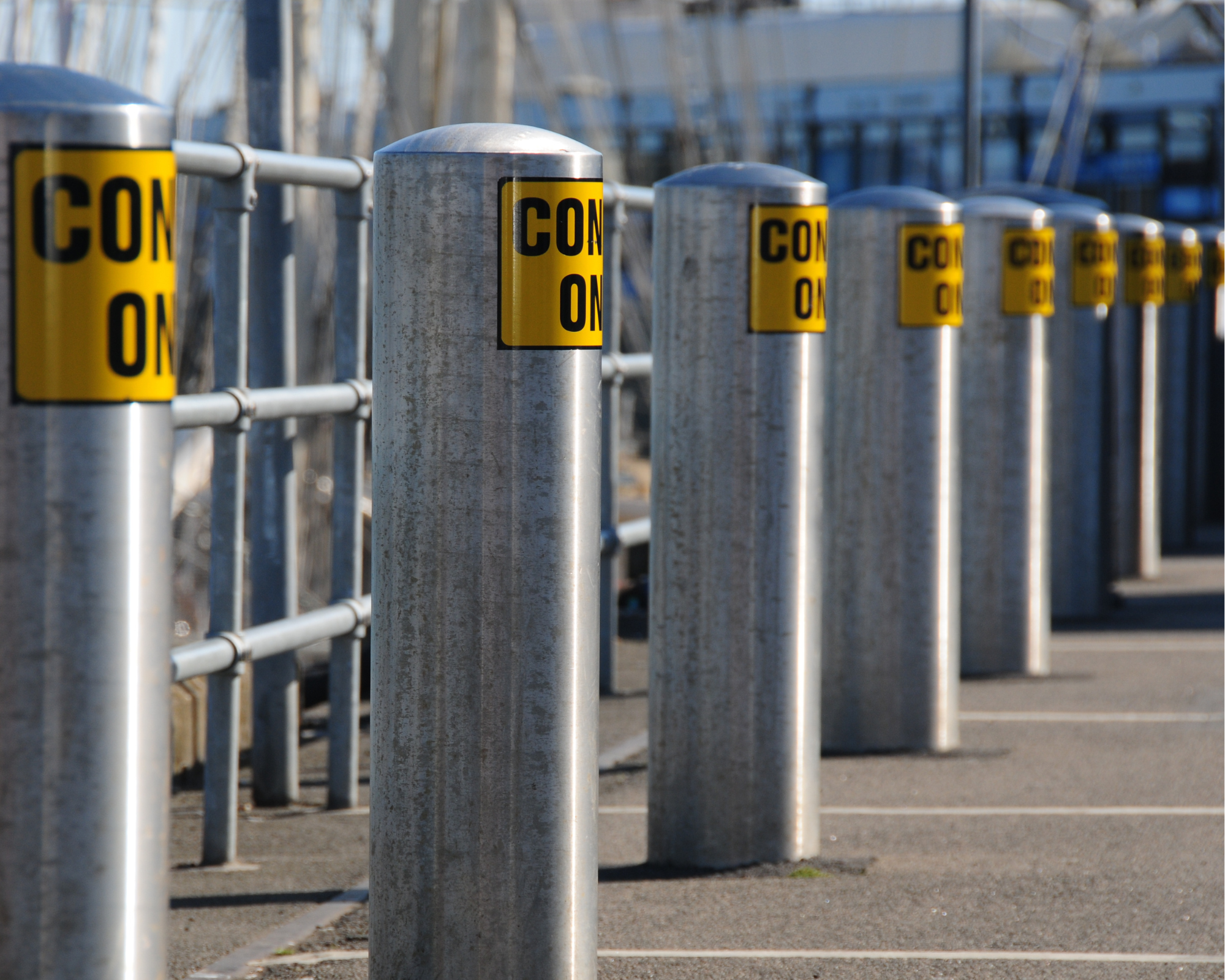 Rangée de bornes de sécurité métalliques avec étiquettes jaunes "CON" visibles, installées le long d’un quai ou d’une voie piétonne.