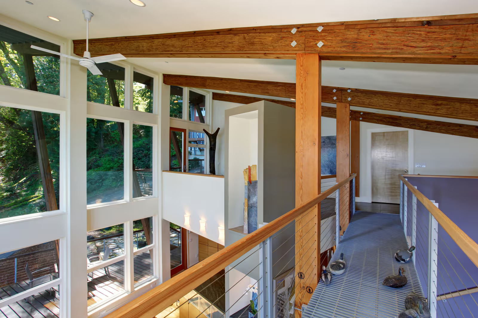 Mezzanine lumineuse avec grandes fenêtres, poutres apparentes en bois, garde-corps en câble métallique et intérieur moderne ouvert sur la nature.