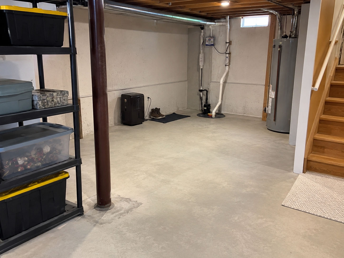 Sous-sol résidentiel sec avec plancher de béton, pompe de puisard, chauffe-eau, étagères de rangement et escalier en bois.