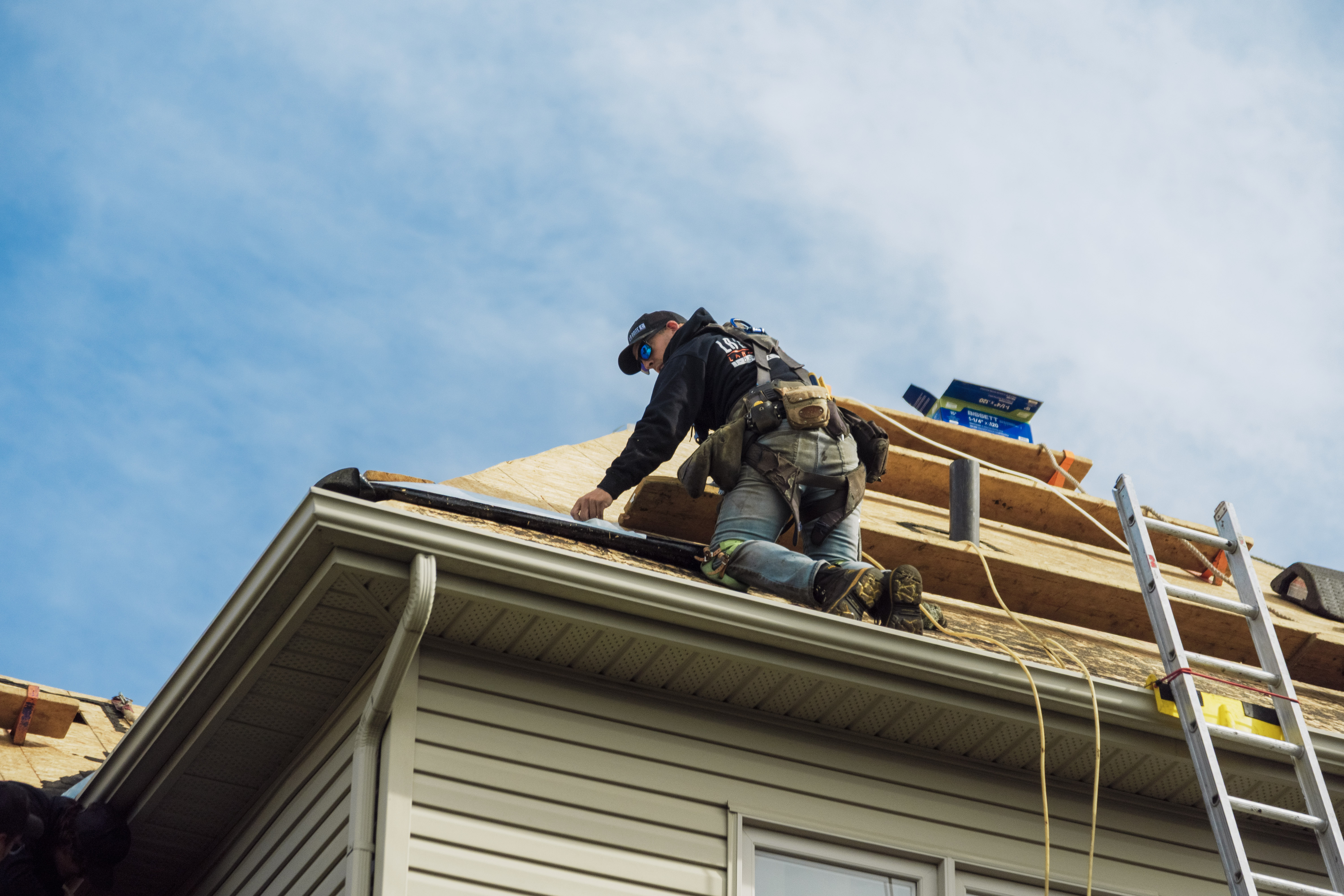 Un ouvrier de construction, équipé de son harnais de sécurité et de lunettes de soleil, installe des panneaux de contreplaqué sur le toit d’une maison, une échelle appuyée contre la façade et des outils posés sur la surface de travail.