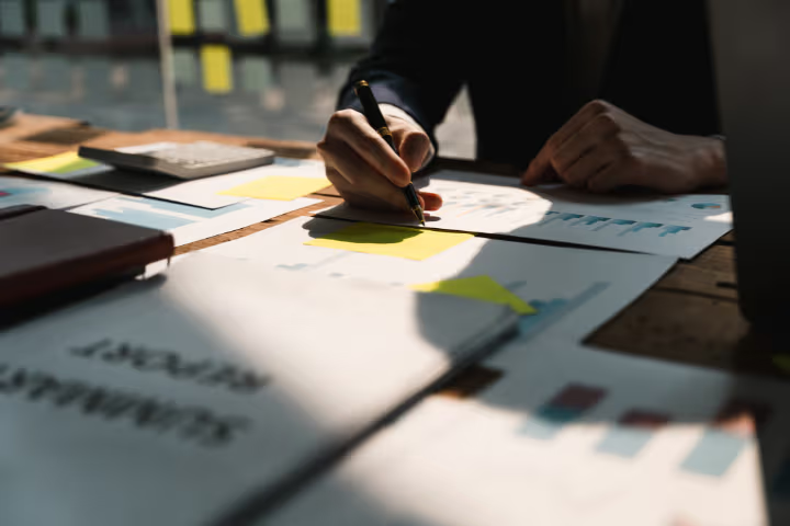 Professional writing on a sticky note among reports, charts and work documents on an office table.