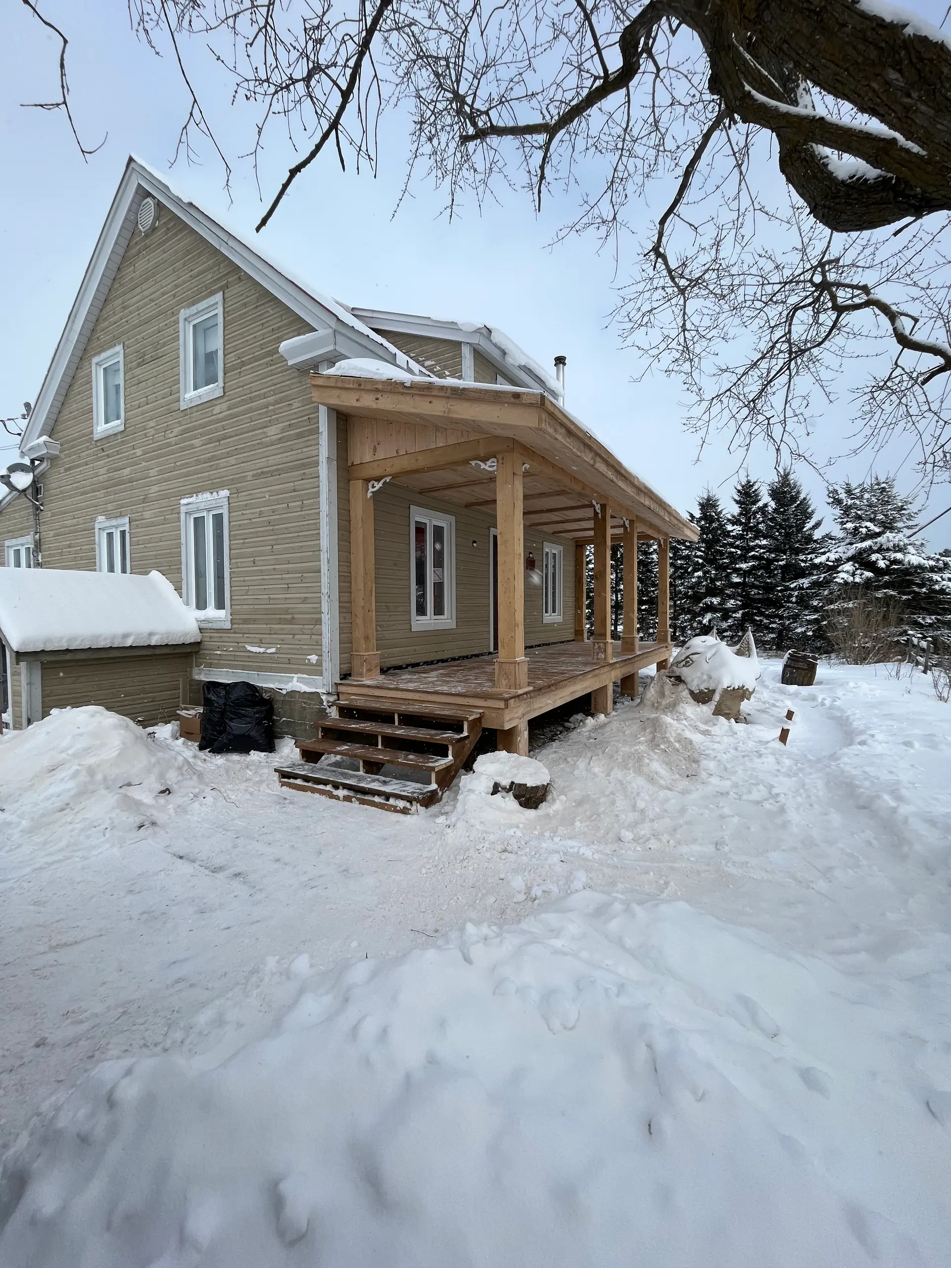 Wooden house with a covered porch under construction in winter, surrounded by snow and evergreen trees.