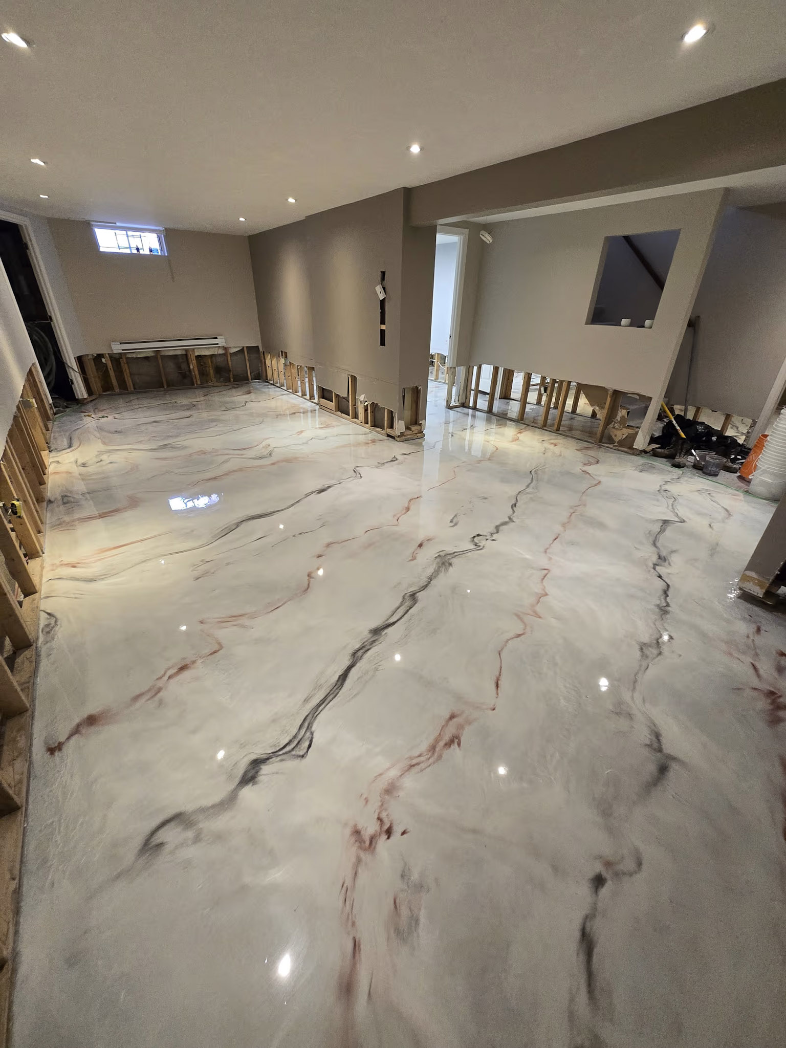 Sous-sol en rénovation avec plancher époxy blanc lustré effet marbre, murs partiellement ouverts, luminaires encastrés et finition brillante
