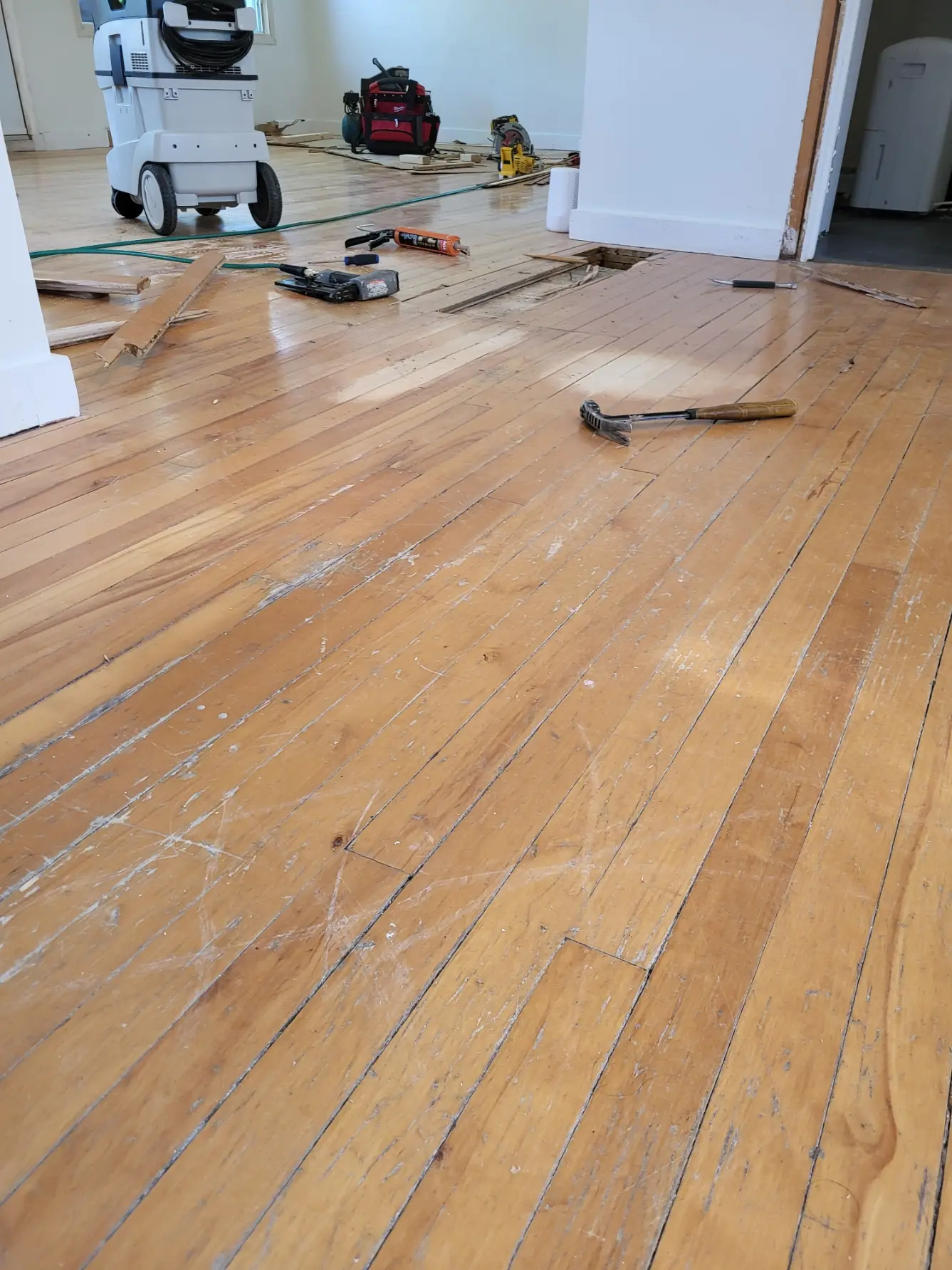 Plancher de bois usé en cours de rénovation, avec outils, cloueuse, marteau et sections de lattes retirées.