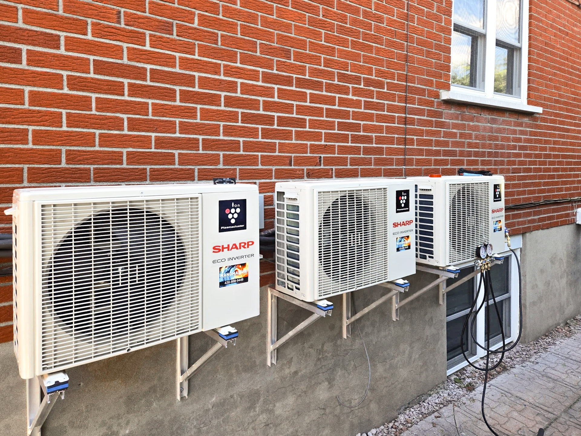 Three Sharp outdoor air conditioning units mounted on a red brick wall of a residential building.