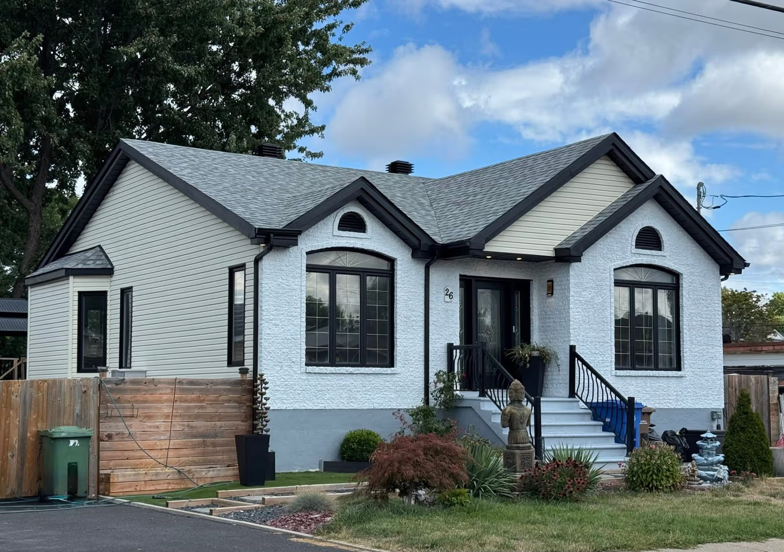 Renovated single-storey house with white stucco façade, beige siding, grey shingle roof, black windows, entrance steps, and landscaped front yard