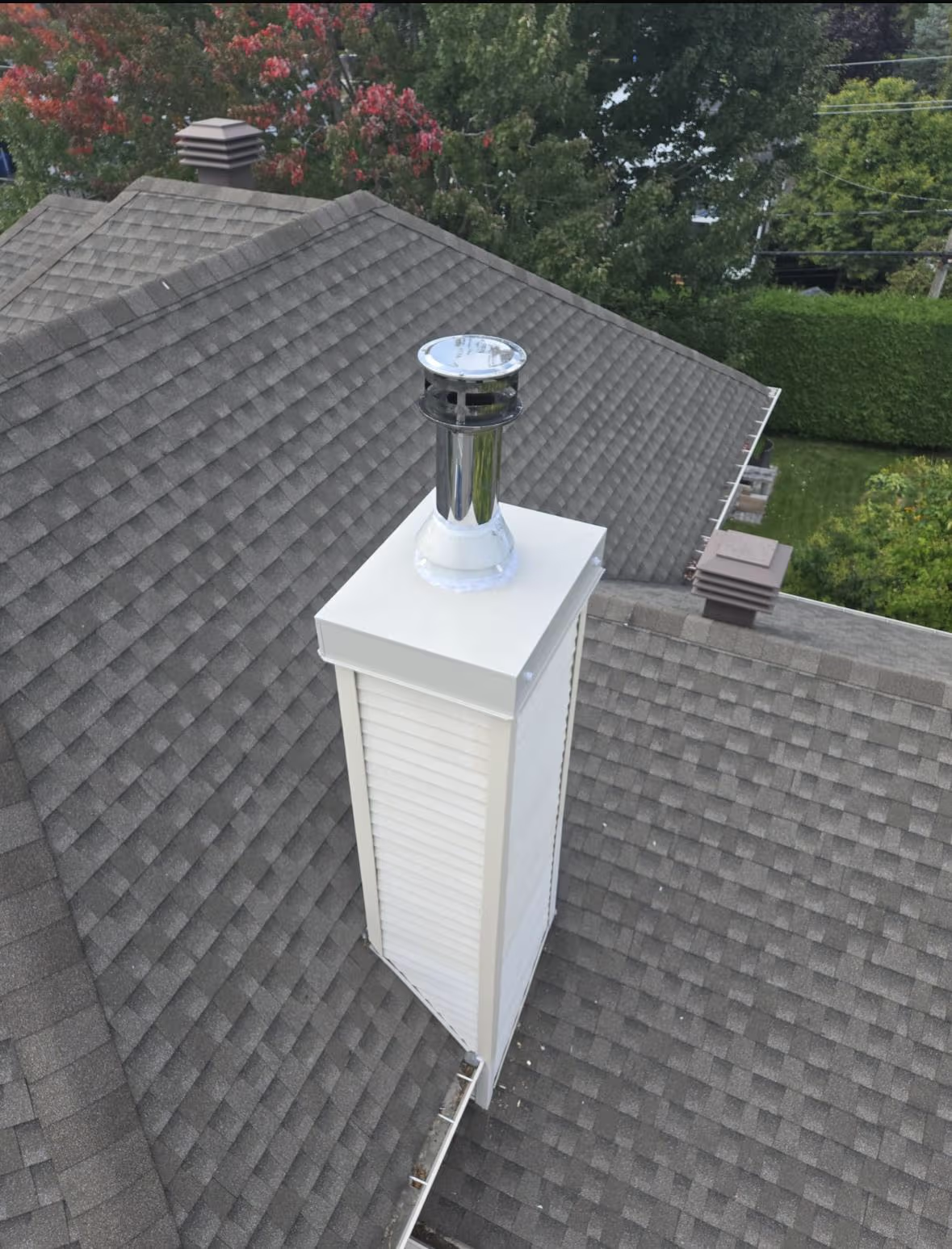 White exterior chimney with metal flue installed on a gray shingle roof surrounded by trees
