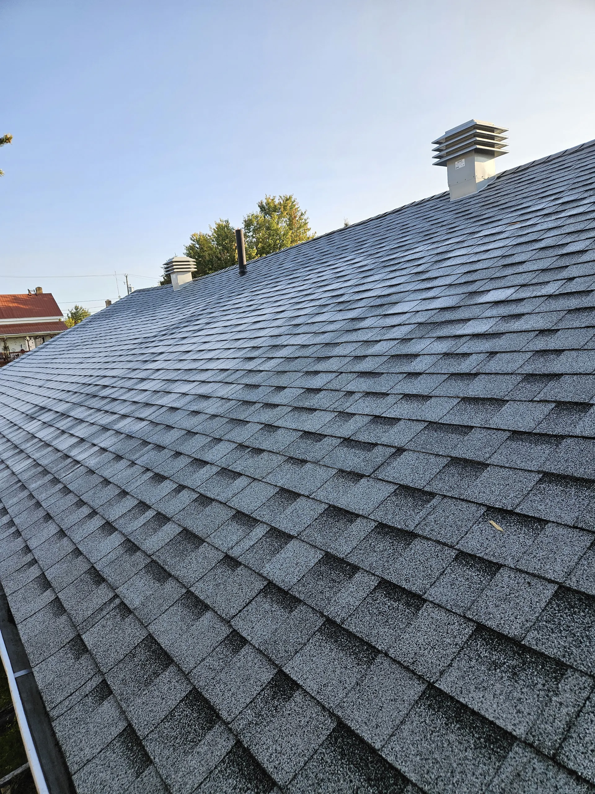 Residential roof covered with gray asphalt shingles with ventilation vents under a clear sky.