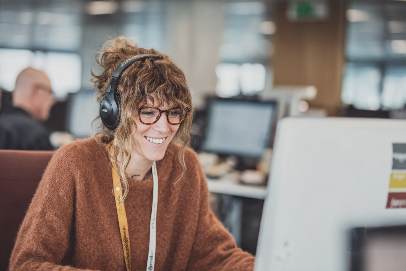 Femme souriante portant des lunettes et un casque audio, travaillant devant un ordinateur dans un bureau à aire ouverte.