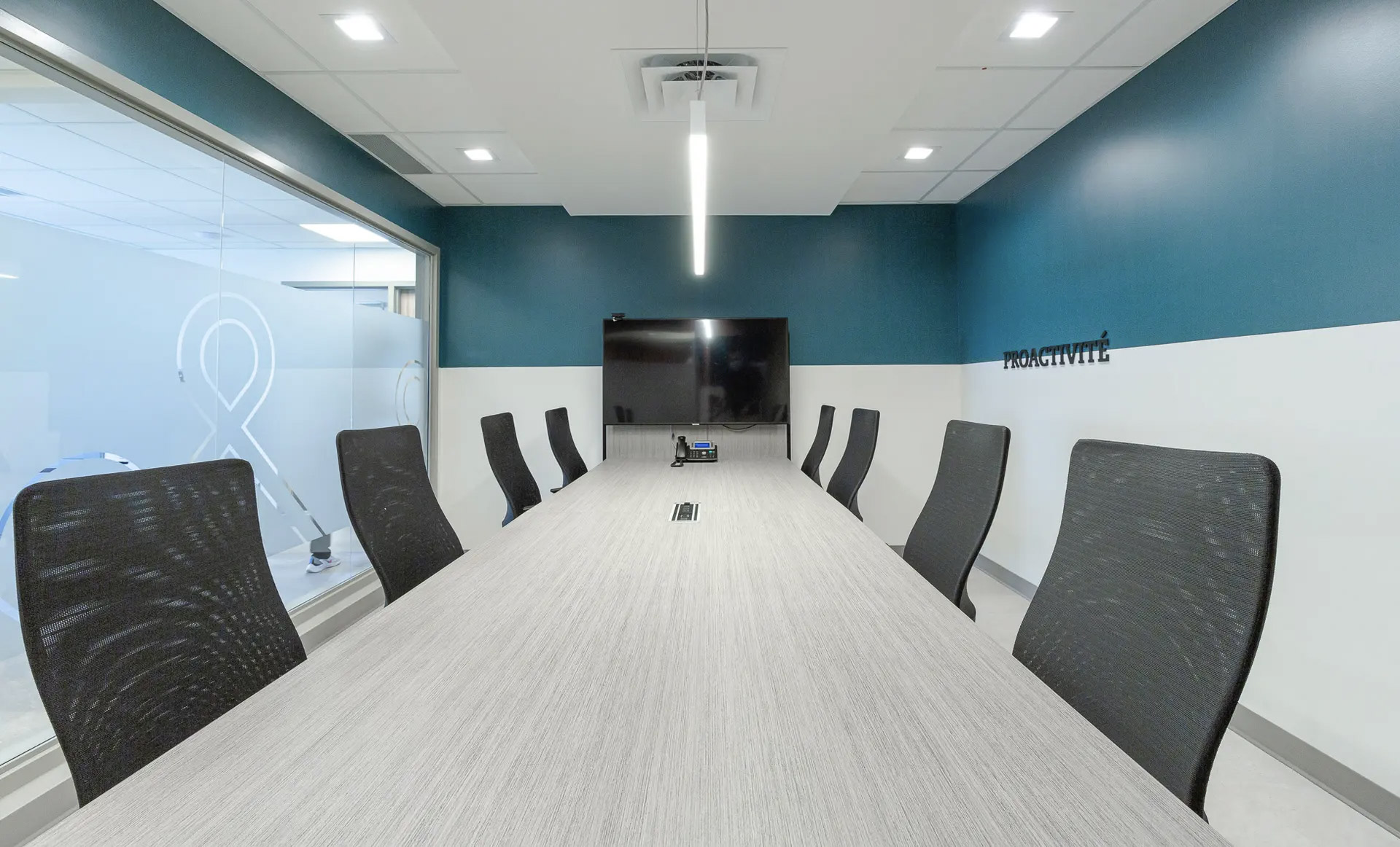 Modern conference room with large table, black chairs, wall screen, glass partition and recessed lighting in a professional office.
