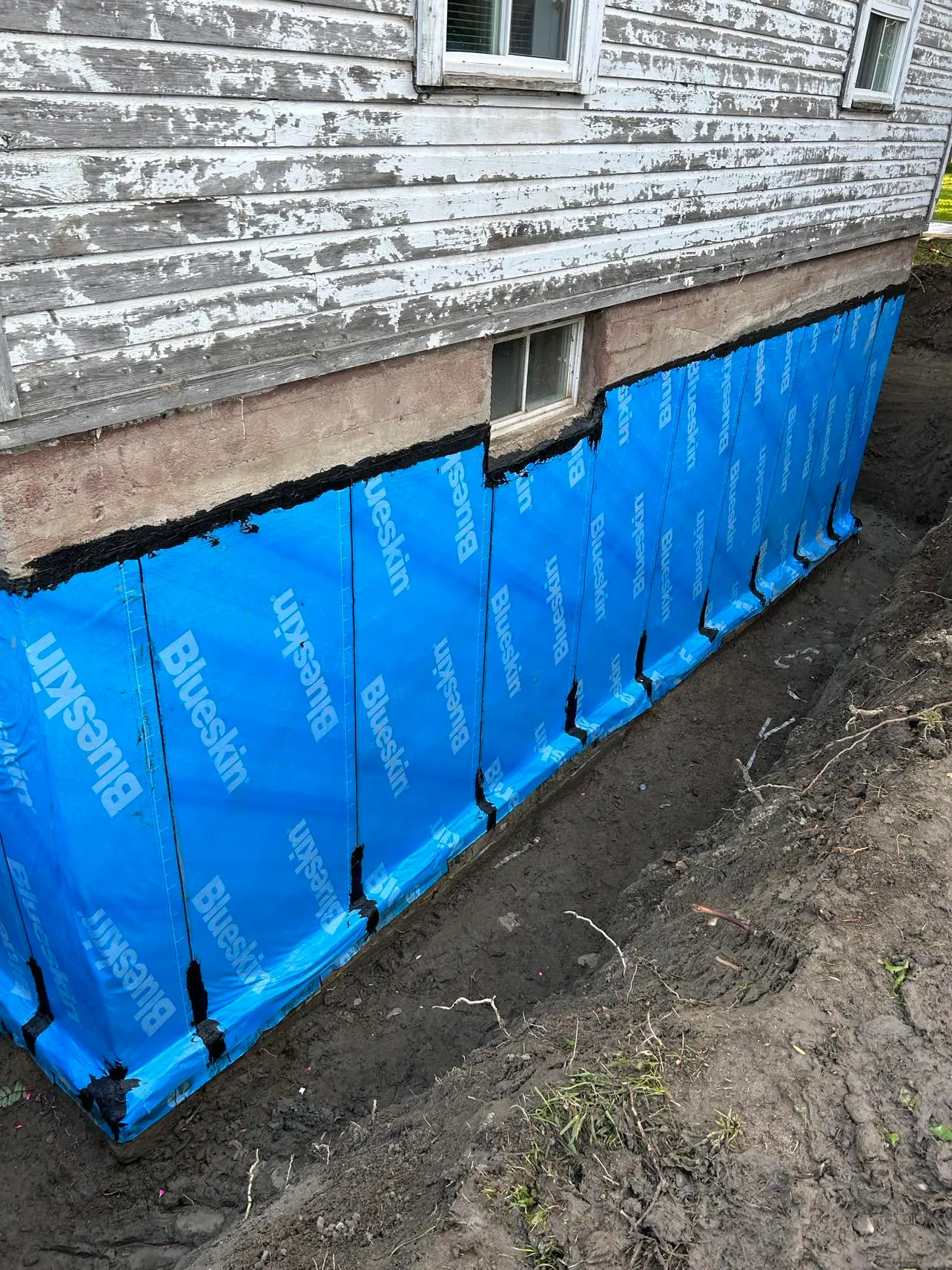 Fondation de maison ancienne avec membrane bleue d’imperméabilisation, excavation extérieure et revêtement de bois écaillé