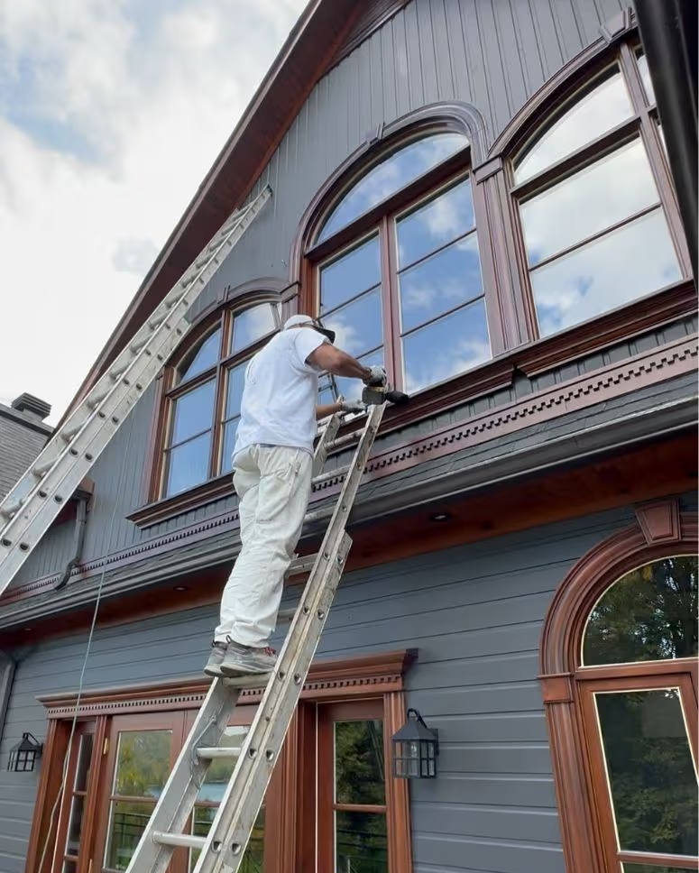 Peintre sur échelle travaillant sur les fenêtres arquées et le revêtement extérieur d’une maison grise