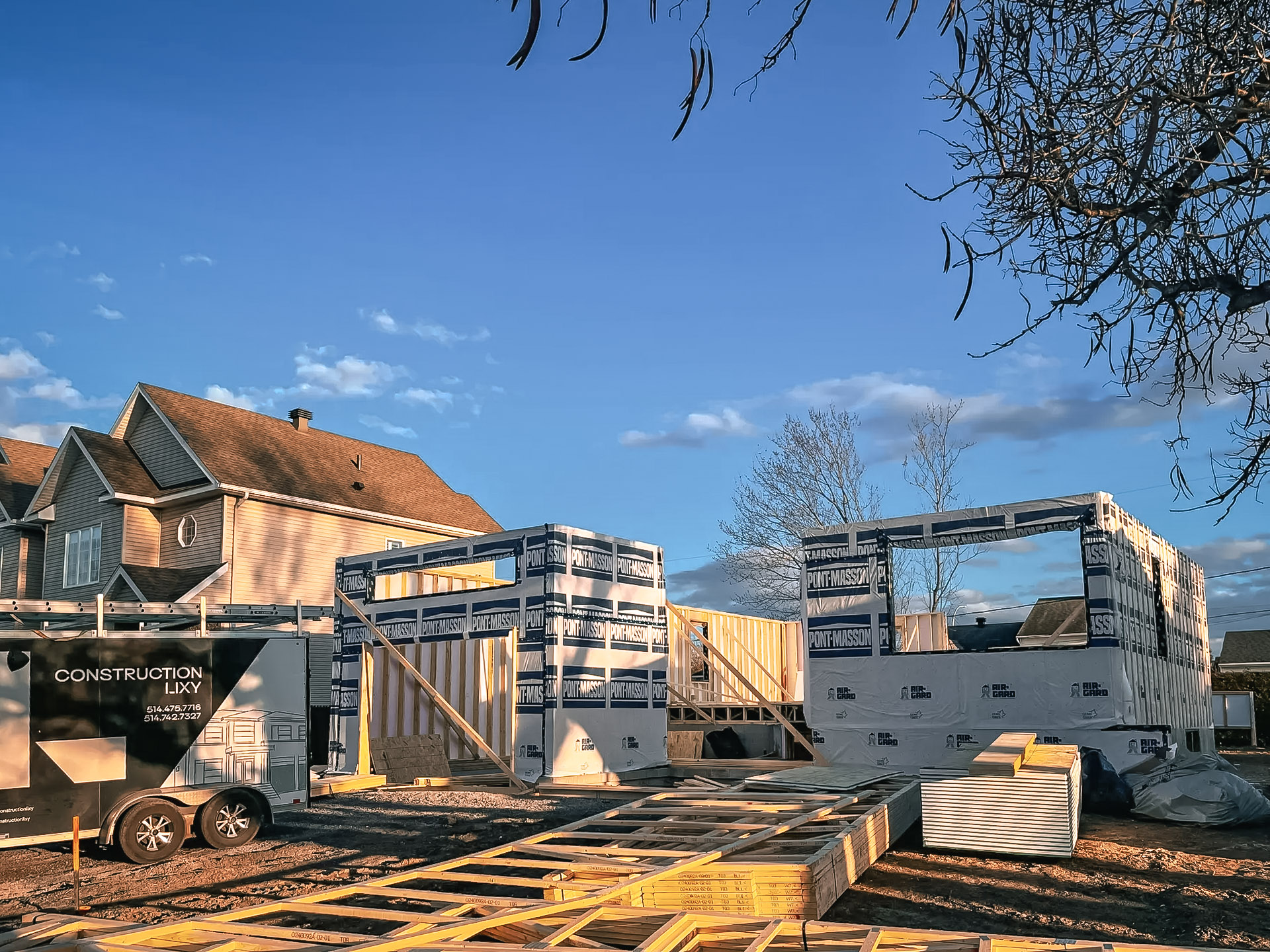 Construction d'une maison en ossature bois avec matériaux empilés et remorque de chantier au coucher du soleil.
