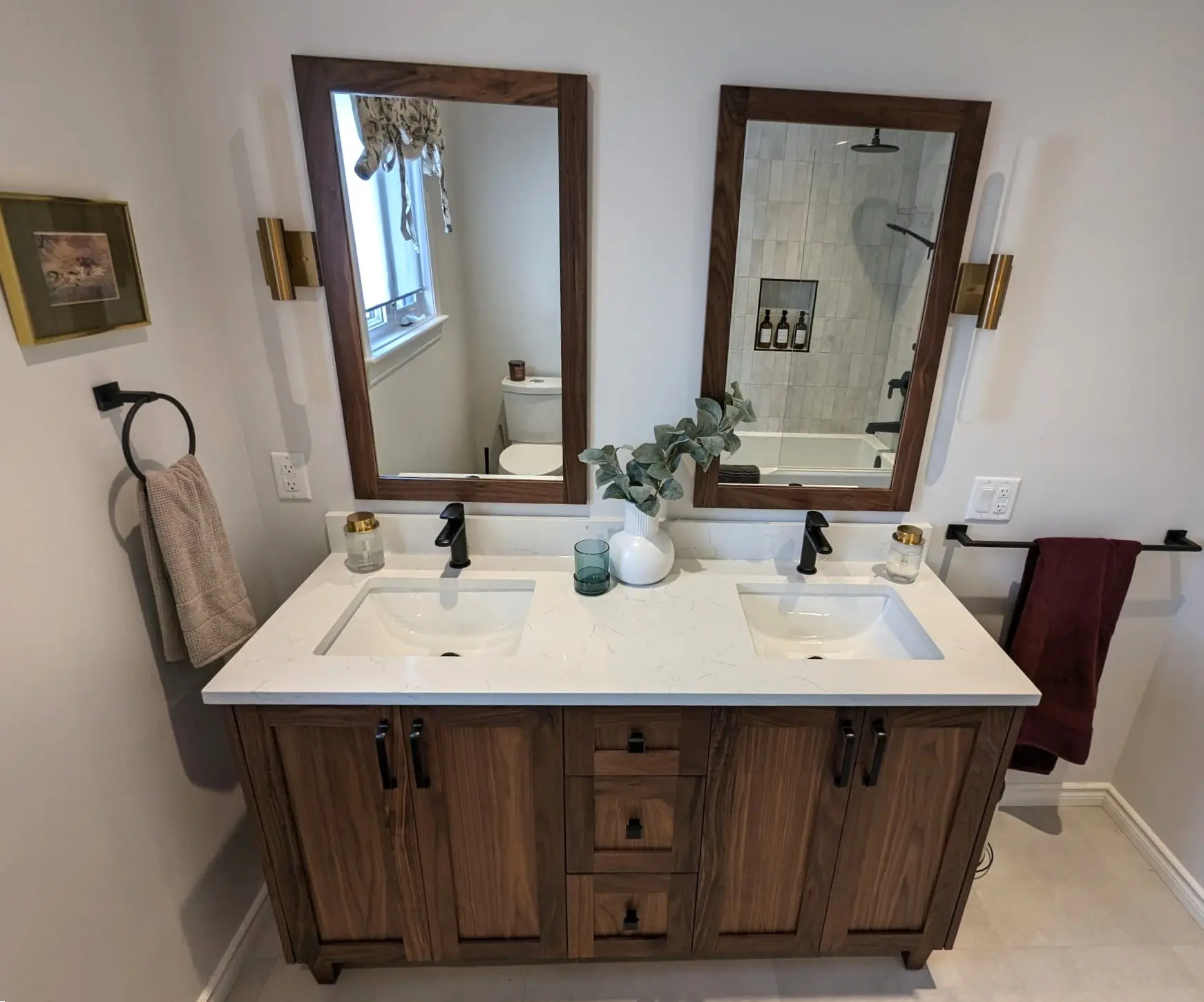 Contemporary bathroom with double wooden vanity, white marble-look countertop, two framed mirrors, black faucets and wall towels