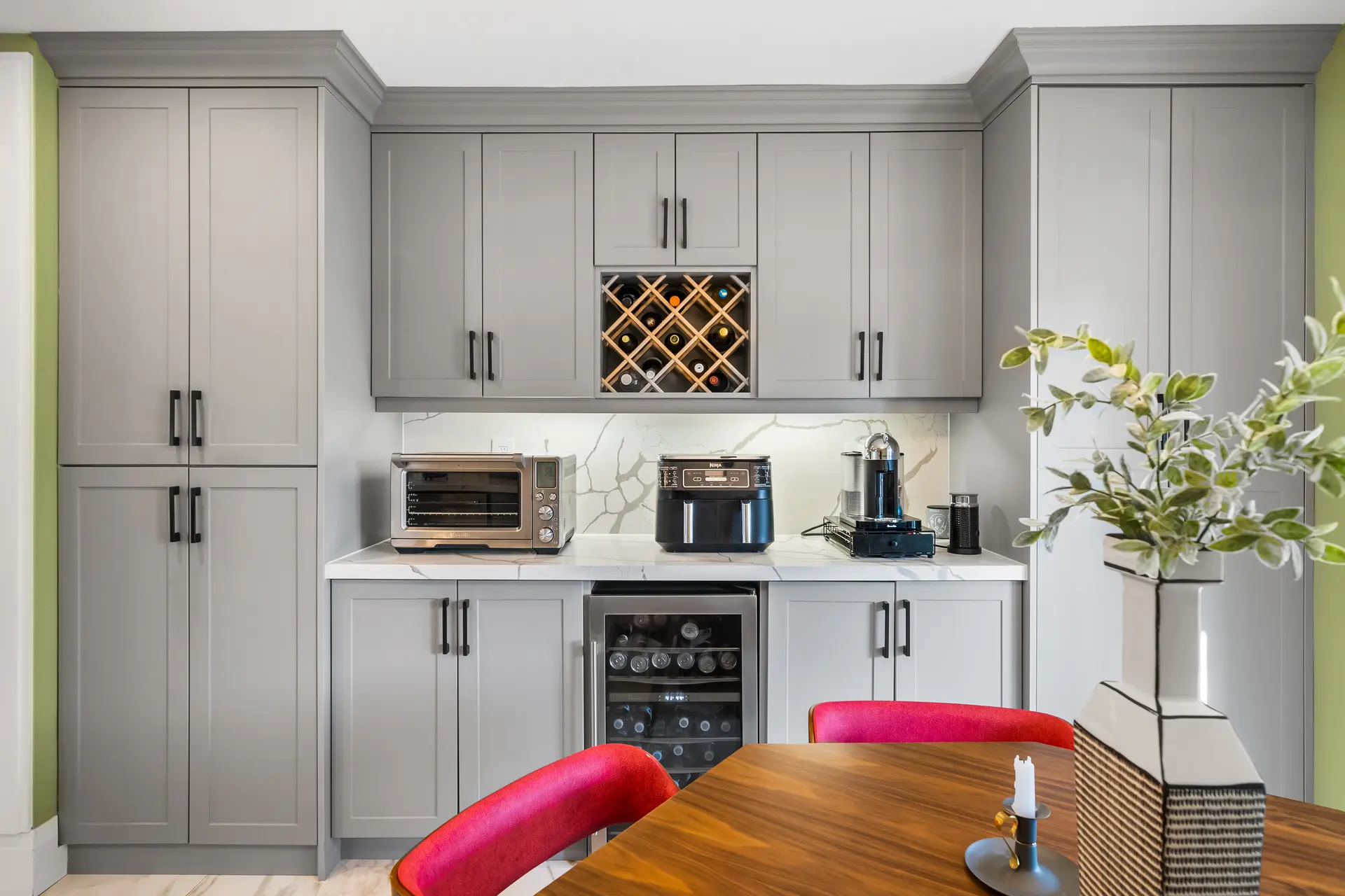 Built-in kitchen with gray cabinets, wine rack, white marble countertop, countertop appliances and wooden table with red chairs