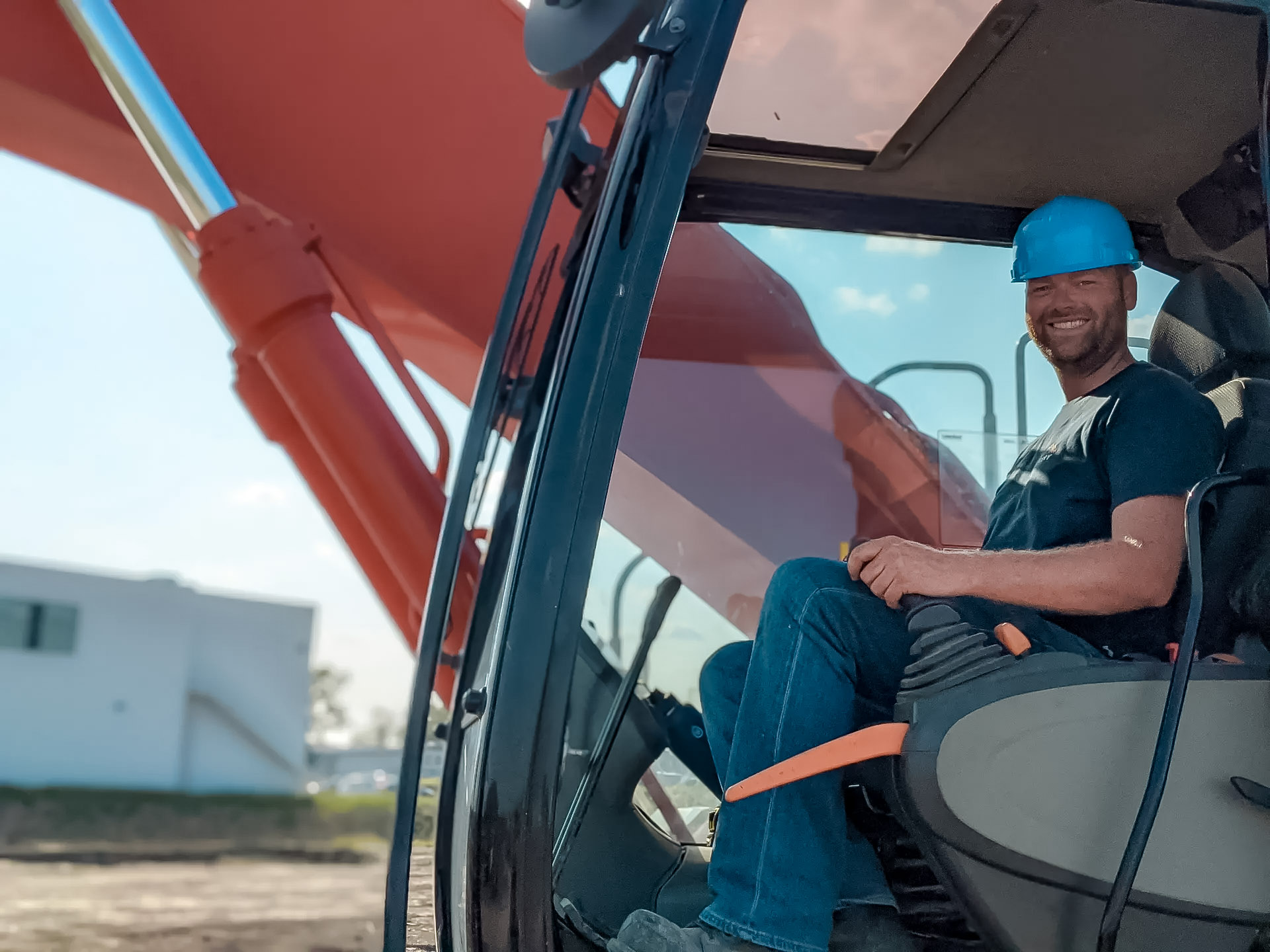 Un travailleur de la construction souriant, portant un casque bleu, est assis dans la cabine d’une excavatrice par une journée ensoleillée sur un chantier.