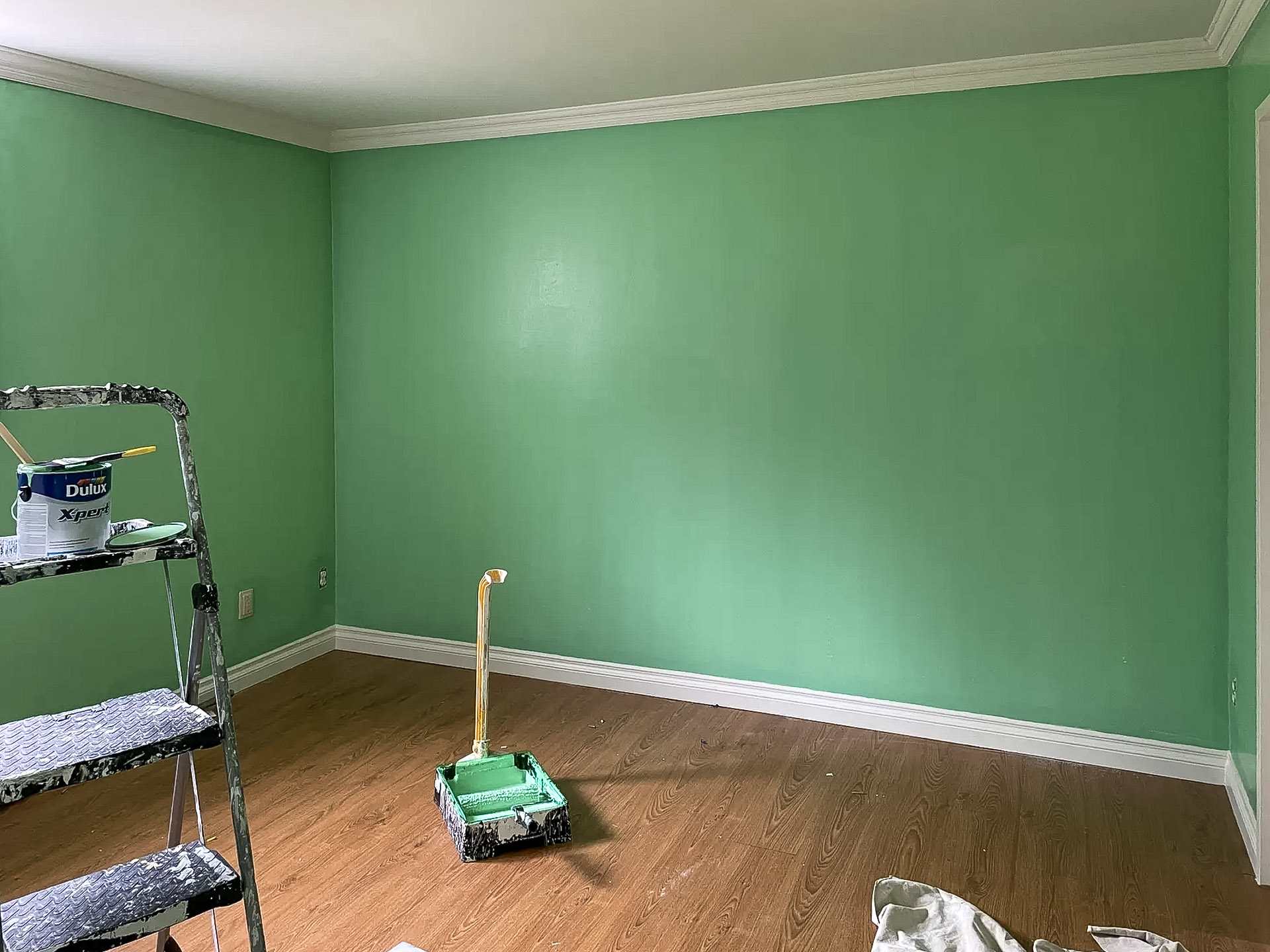 Chambre en cours de rénovation avec murs fraîchement peints en vert, moulures blanches, sol en bois et matériel de peinture au sol.