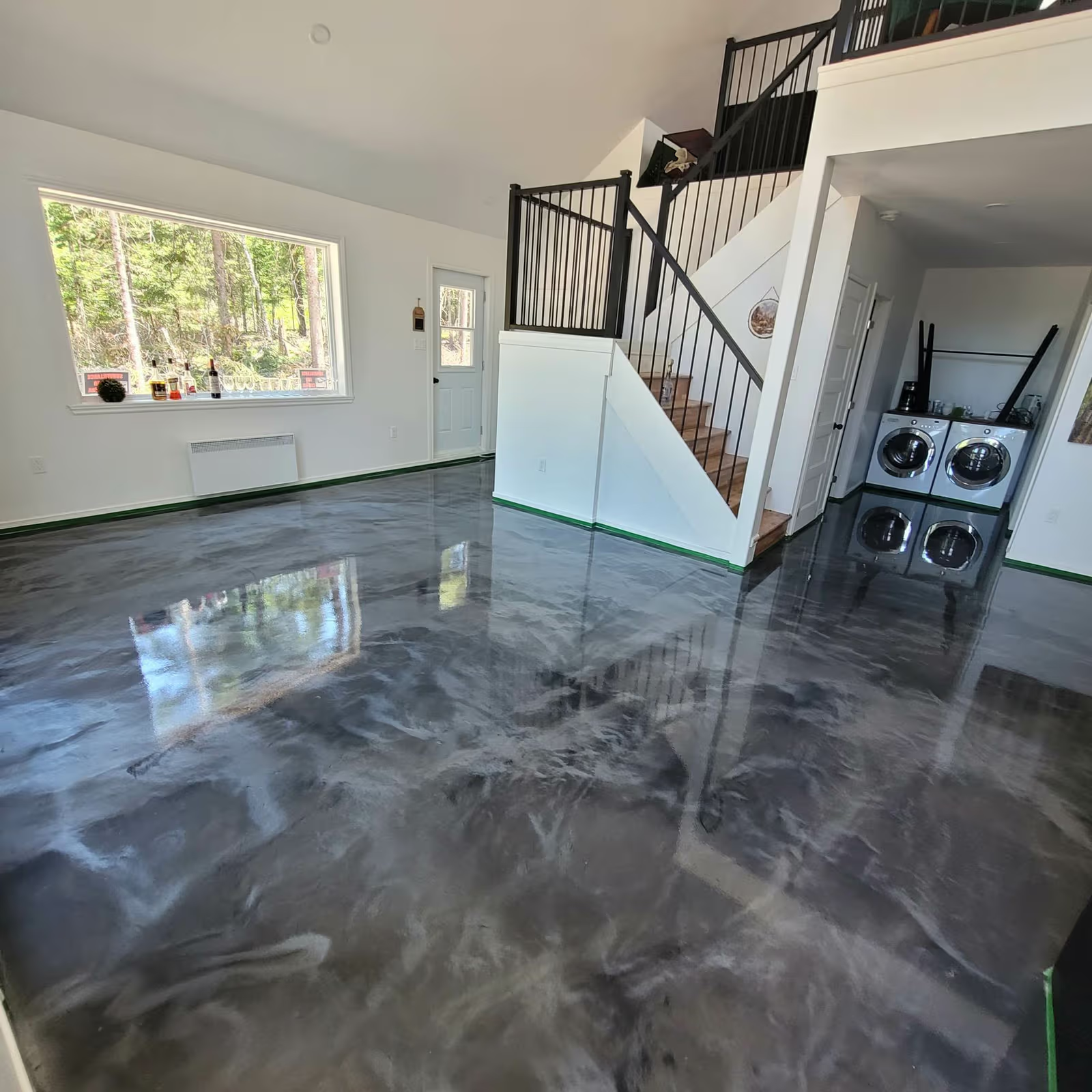 Intérieur résidentiel moderne avec plancher époxy gris lustré, escalier en bois, rampes noires, murs blancs, grande fenêtre et coin buanderie