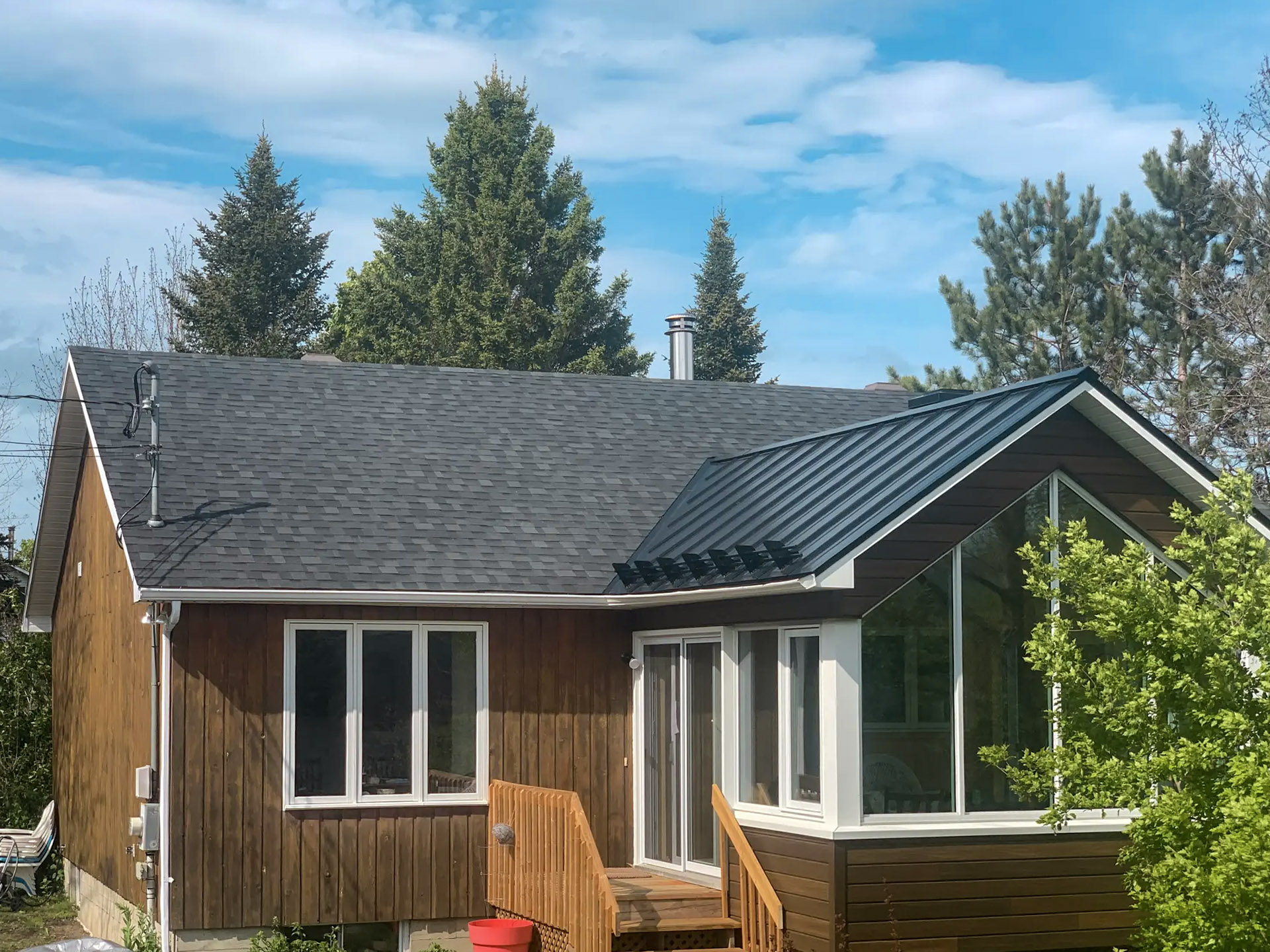 Typical Montreal suburban bungalow with wood siding, mixed asphalt shingle and black sheet metal roof, glass extension and exterior wooden staircase.