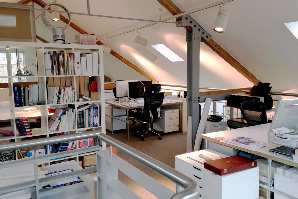 Bureau en mezzanine sous plafond incliné avec poutres apparentes en bois, puits de lumière, étagère de livres, postes de travail et garde-corps métallique.