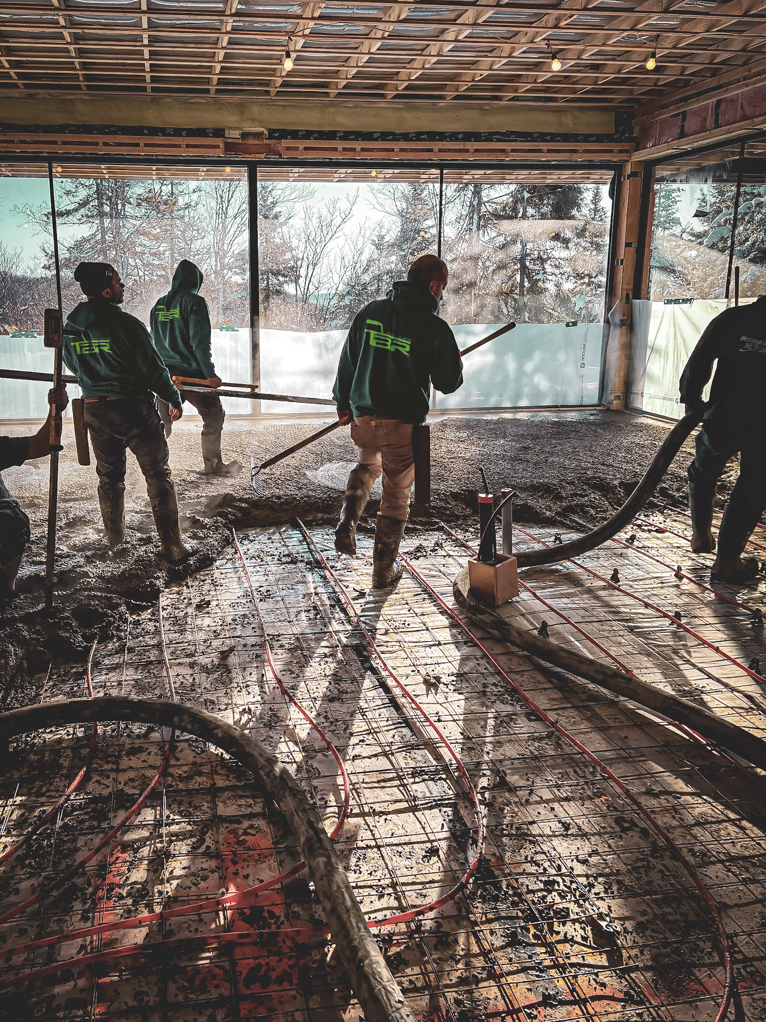 Crew of workers pouring a concrete slab over a red-pipe radiant floor heating system in a building under construction.