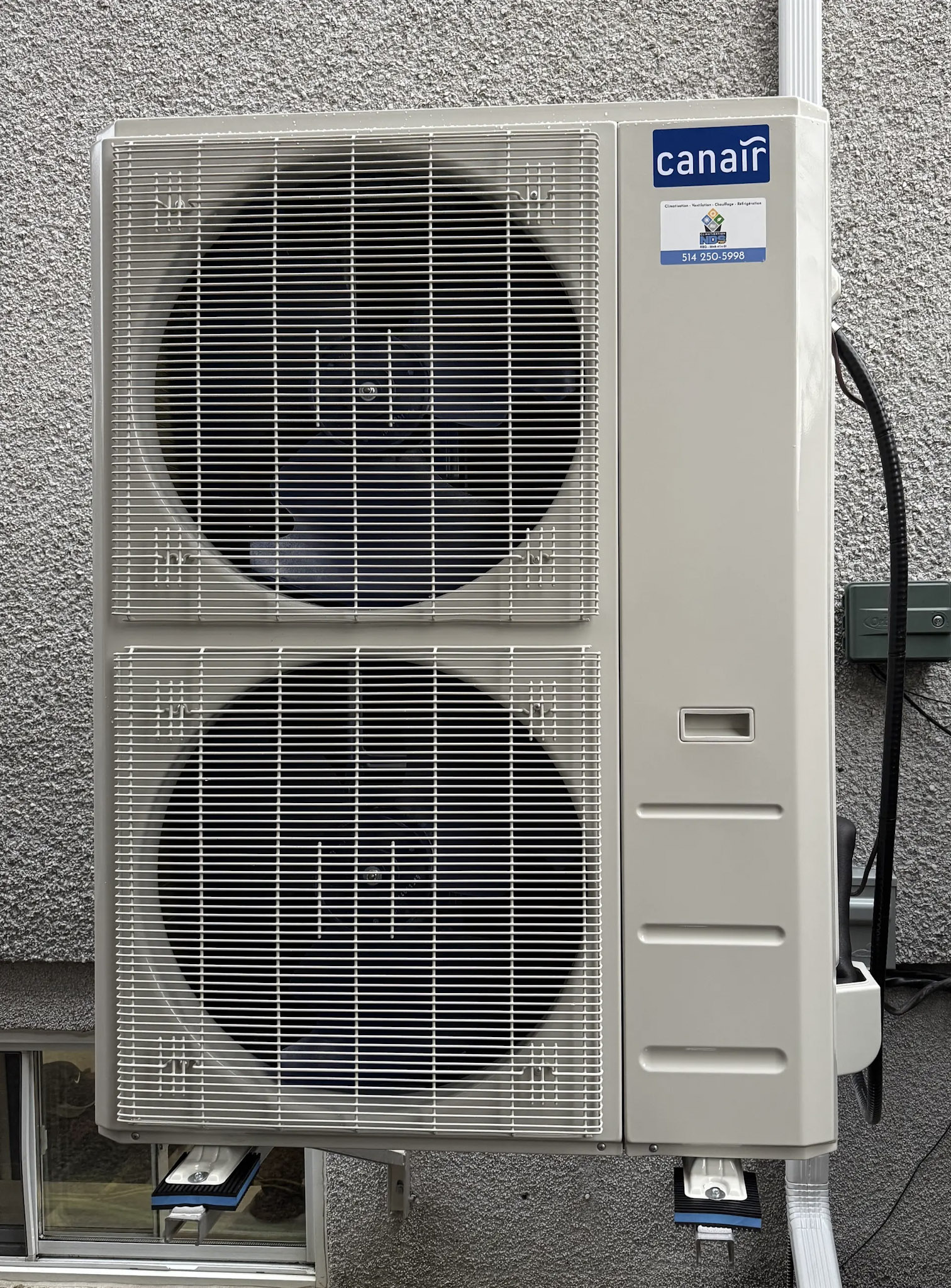 Unité extérieure de climatisation ou thermopompe murale avec deux ventilateurs, grilles blanches, câblage apparent et fixation sur mur en crépi gris