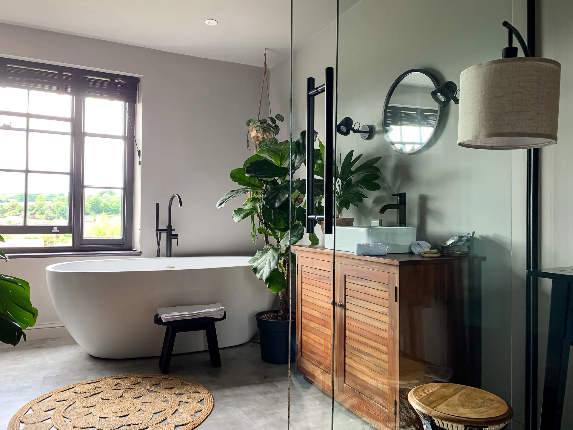 Salle de bain avec baignoire îlot blanche, meuble en bois naturel, plantes vertes, miroir rond et éclairage moderne.