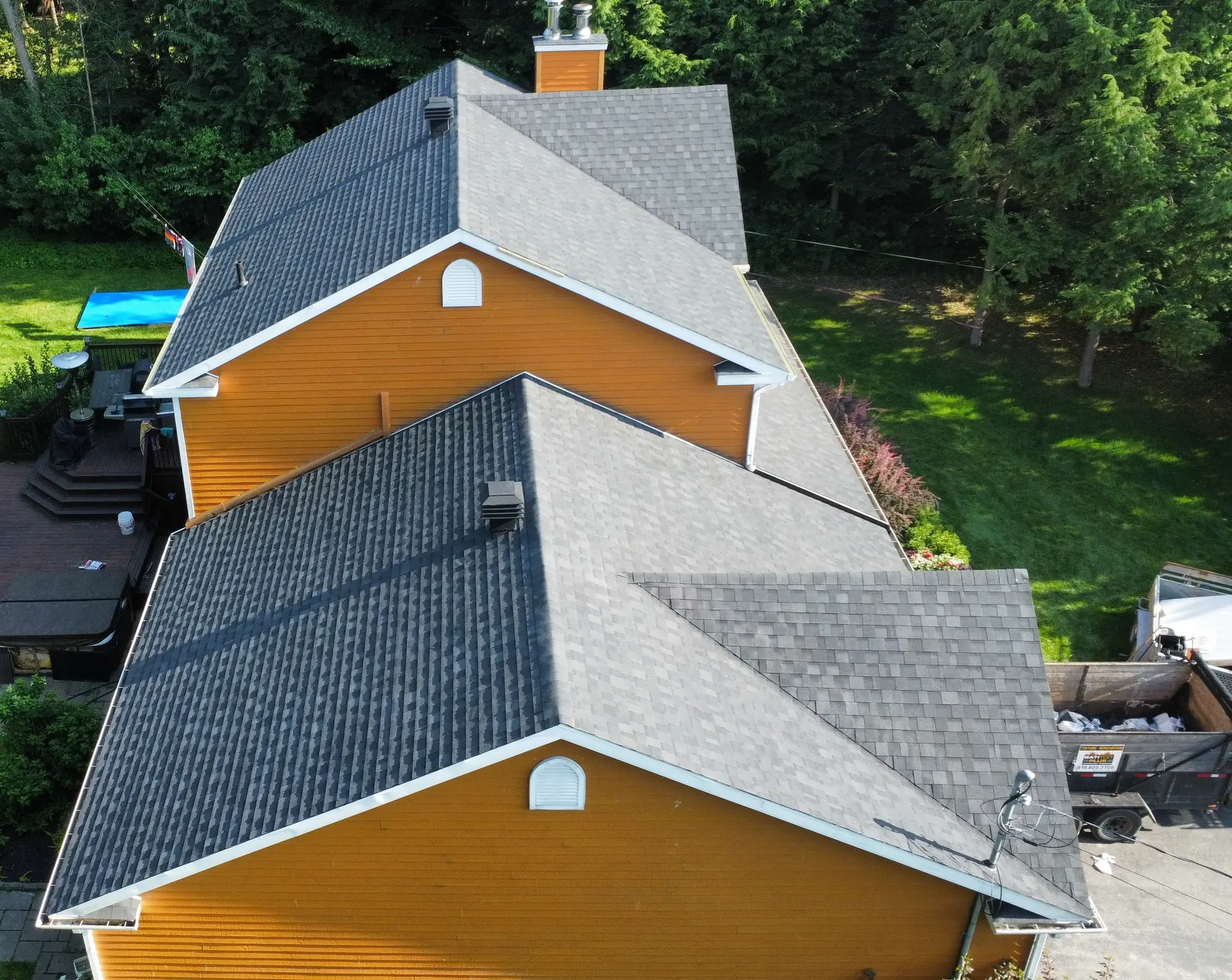 Aerial view of a yellow house with grey shingle roofing, multiple gables, roof vents, and a wooded yard.