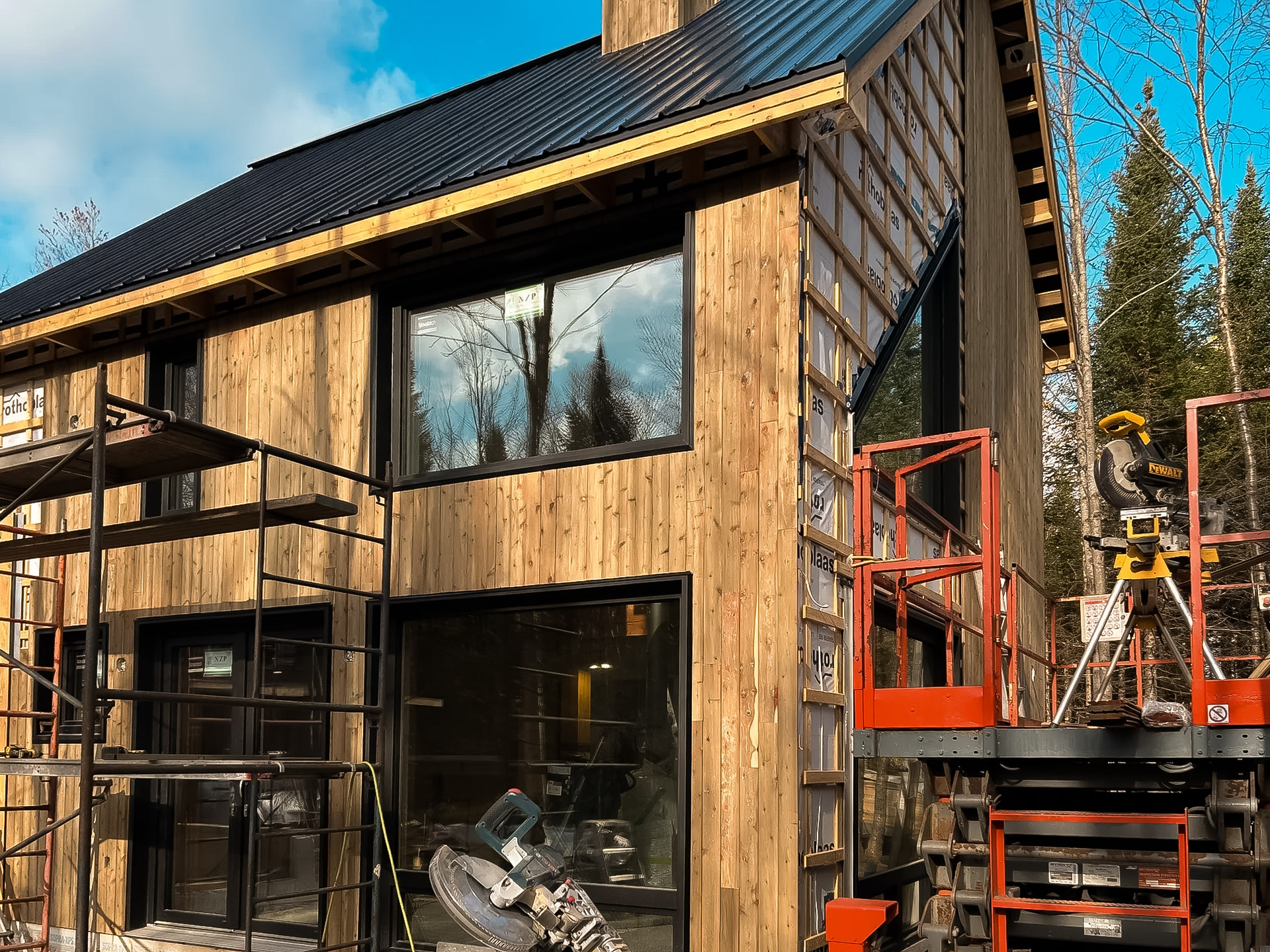 House under construction with wooden exterior cladding, large windows and black tin roof, surrounded by scaffolding and tools.
