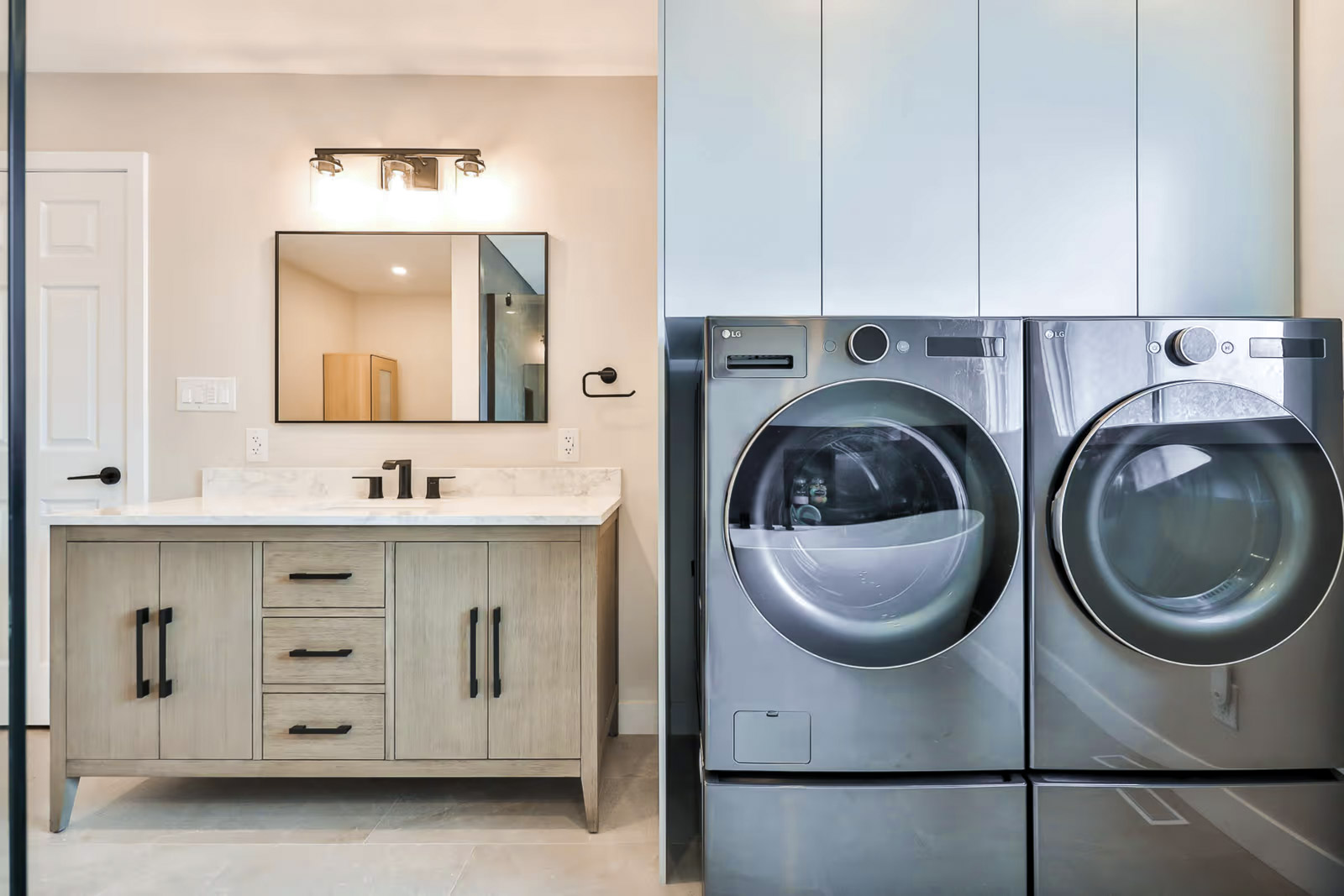 Modern bathroom with integrated laundry area, gray washer and dryer, light wood vanity, white countertop, wall mirror, black fixtures, wall lighting and beige ceramic floor for a bright functional space