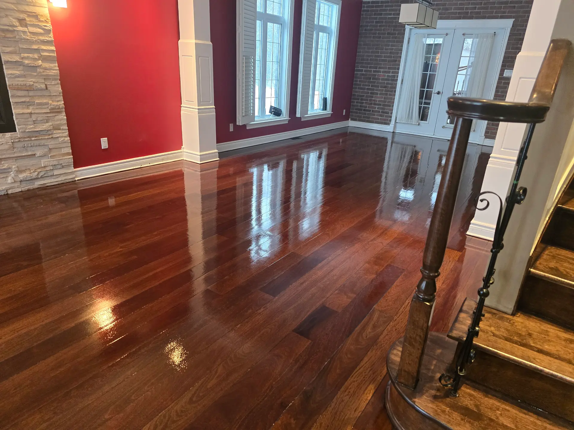 Freshly varnished dark brown hardwood floor in a bright large room with red walls, a stone fireplace, and staircase.