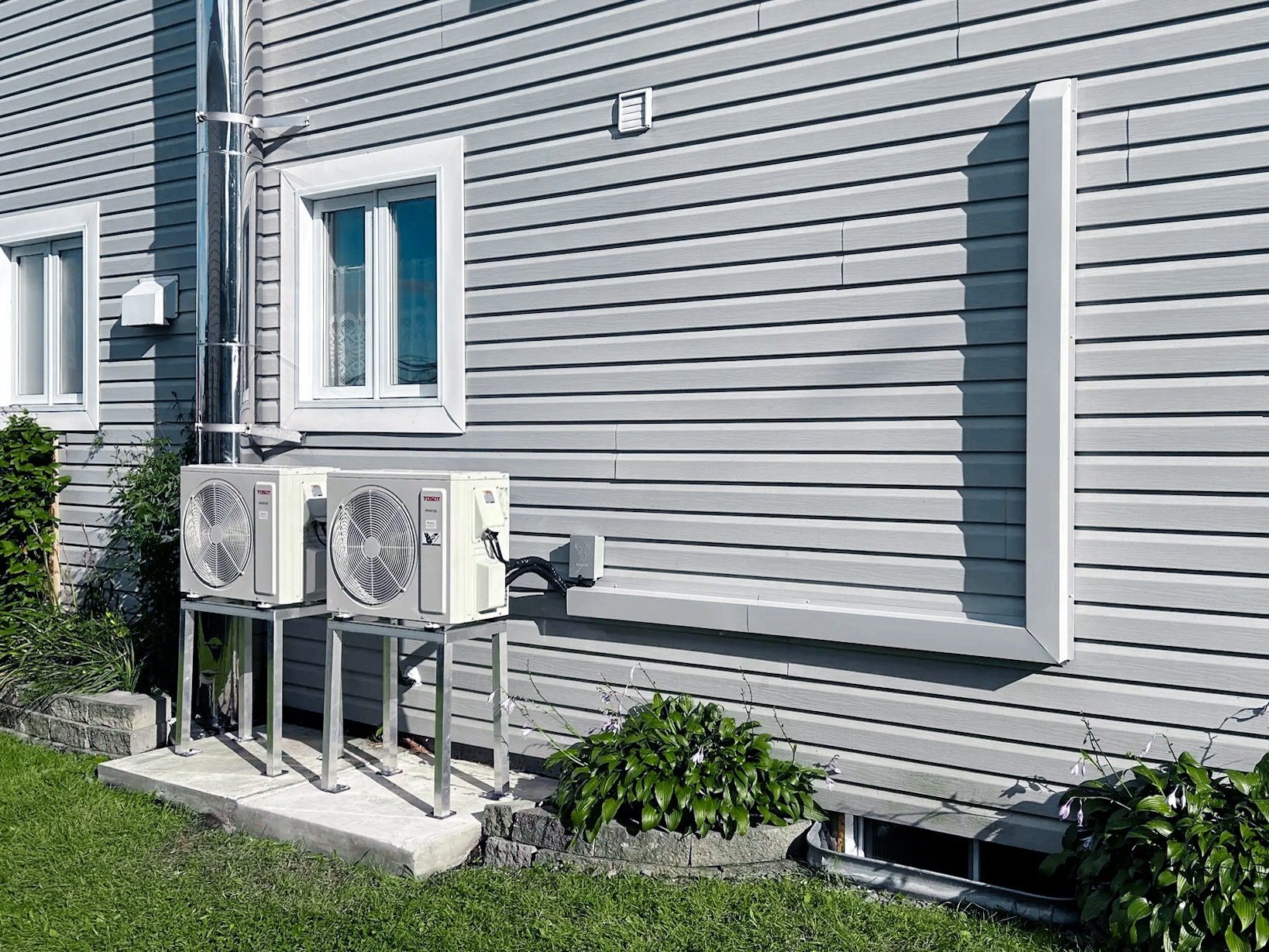 Two Tosot outdoor air conditioning units mounted on metal stands along a grey vinyl-sided house wall.