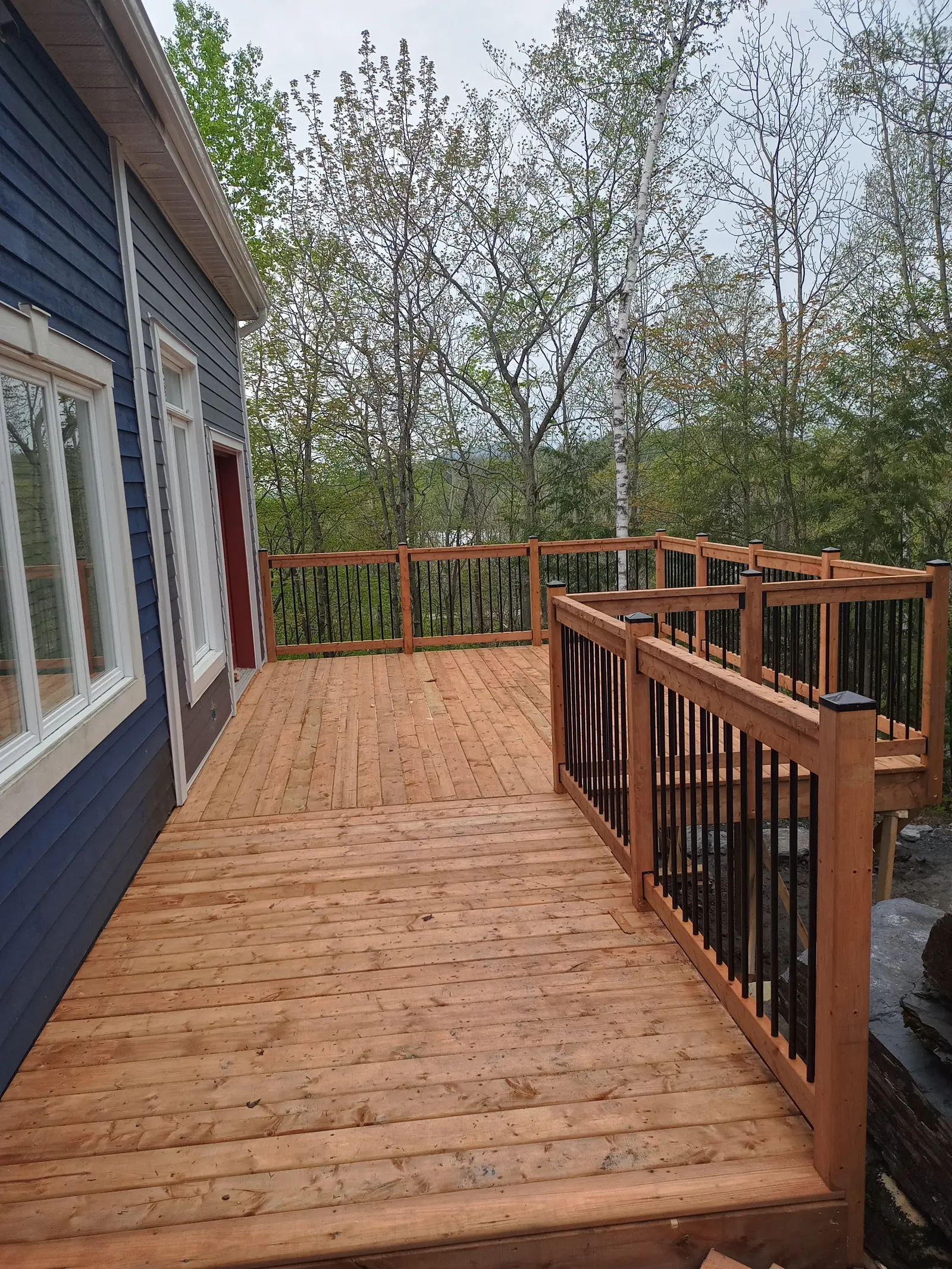 Grande terrasse en bois avec garde-corps noir et bois, attenante à une maison bleue en bordure de forêt
