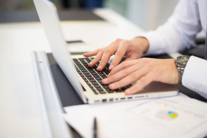 Mains d’un professionnel tapant sur un ordinateur portable avec des documents de travail sur un bureau moderne.
