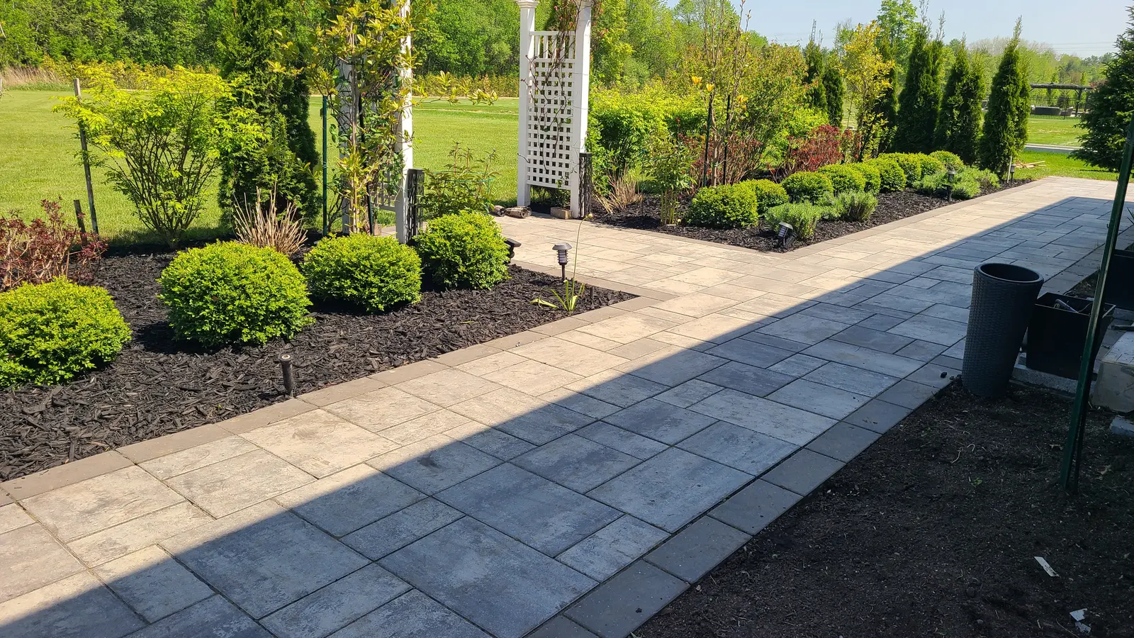 Garden walkway with grey interlocking pavers, borders, landscaped flower beds, shrubs, a white trellis, and outdoor lighting.