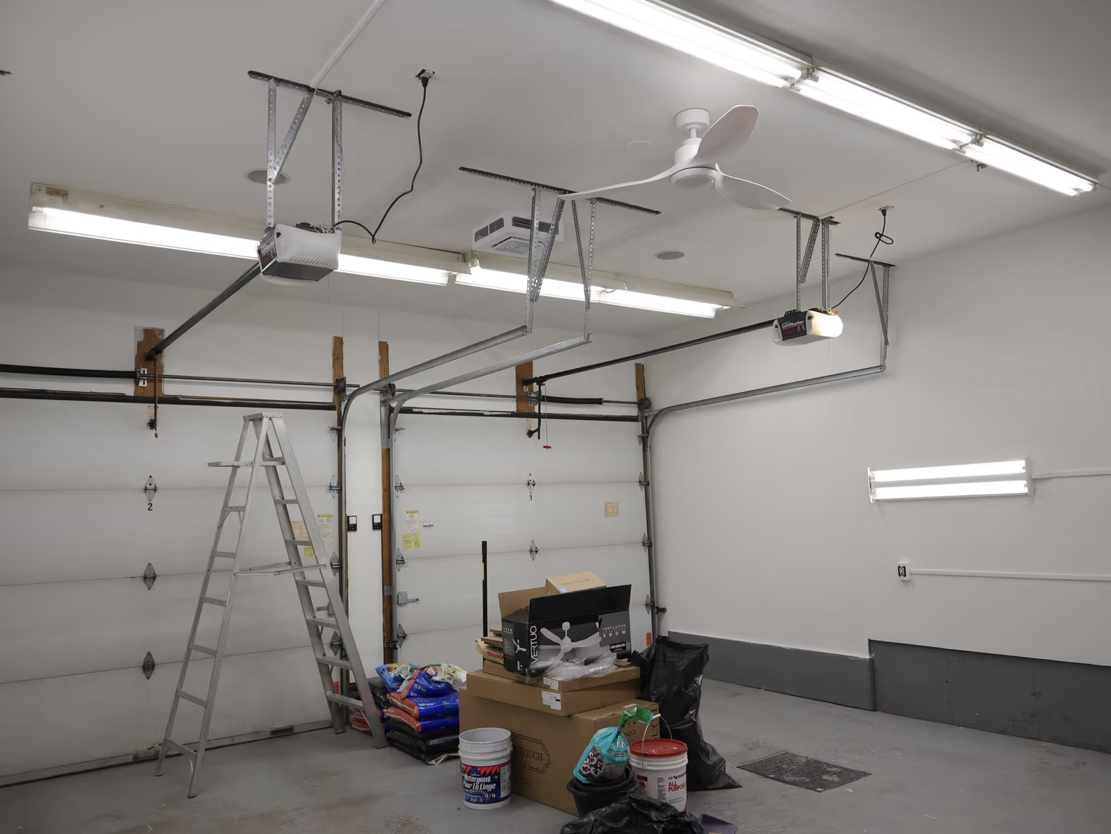 Interior of a double garage with sectional doors, motorized garage door openers, fluorescent lighting, ceiling fan, ladder, and stored materials.