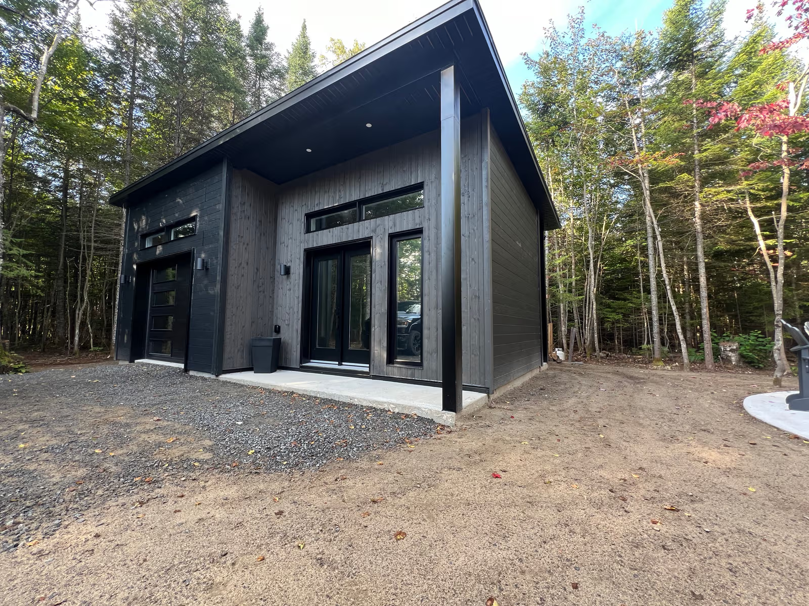 Maison contemporaine en forêt avec revêtement extérieur noir et bois gris, porte de garage moderne, grandes portes vitrées et dalle de béton