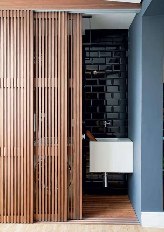 Salle de bain moderne avec cloison coulissante en lattes de bois, lavabo blanc, carrelage noir brillant et murs bleus.