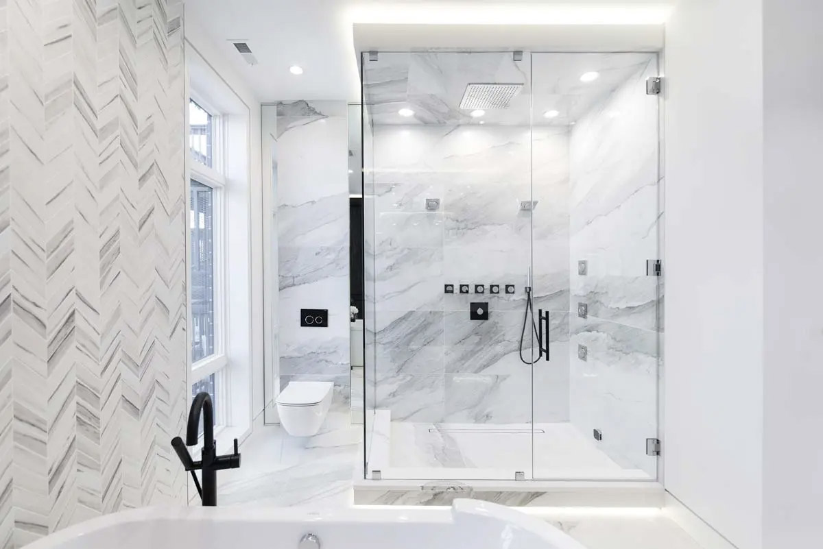 Modern and luxurious bathroom with glass shower, white marble walls, chevron tiles, black faucets, white bathtub and recessed lighting for a sleek and bright design