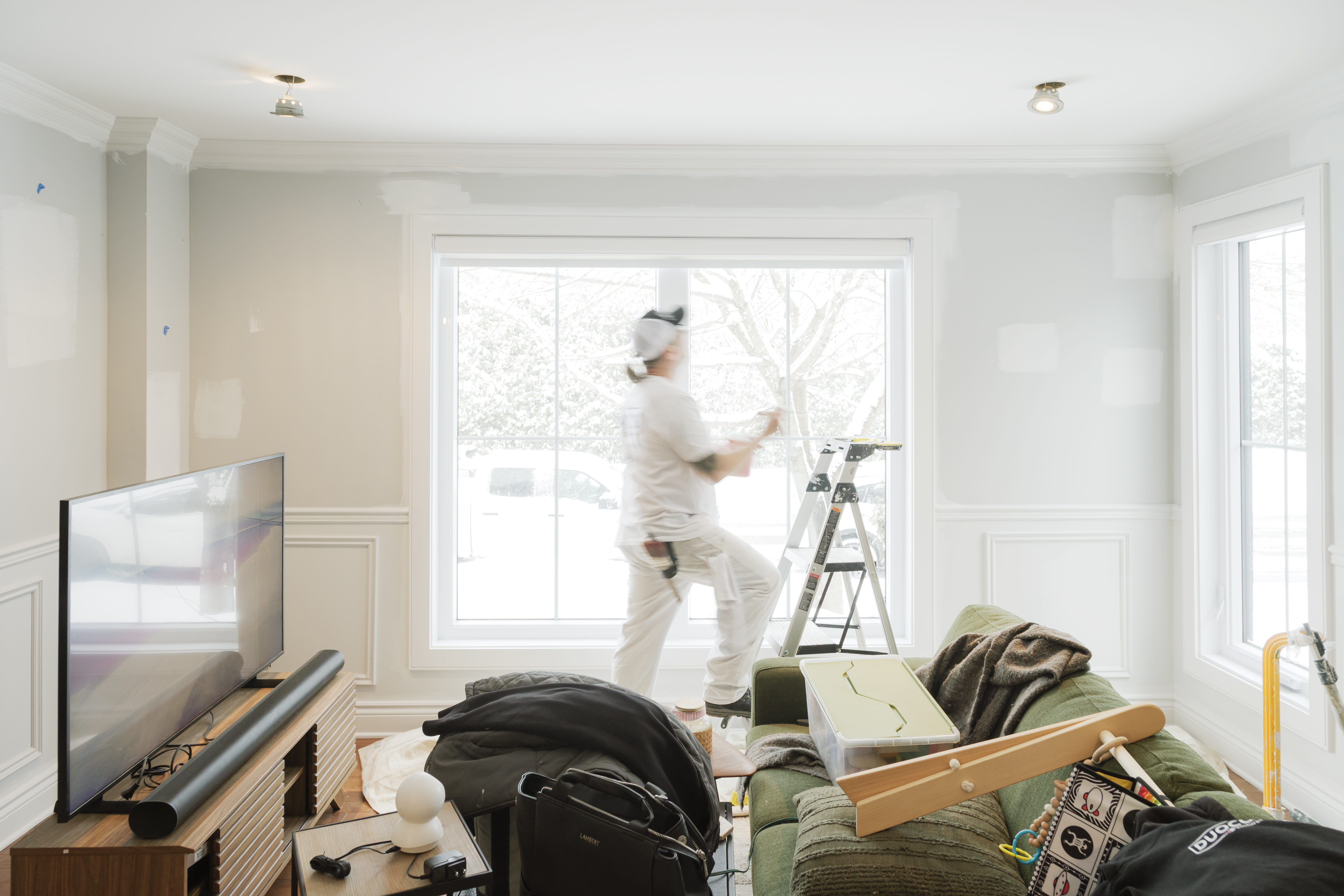 Professional painter renovating the walls of a bright living room with moldings, large windows, step ladder, and protected furniture