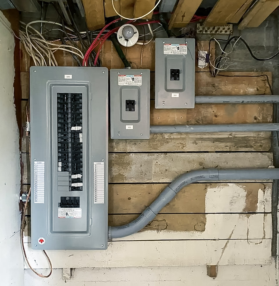 Panneau électrique principal et sous-panneaux installés sur un mur en bois brut avec conduits gris et câblage apparent